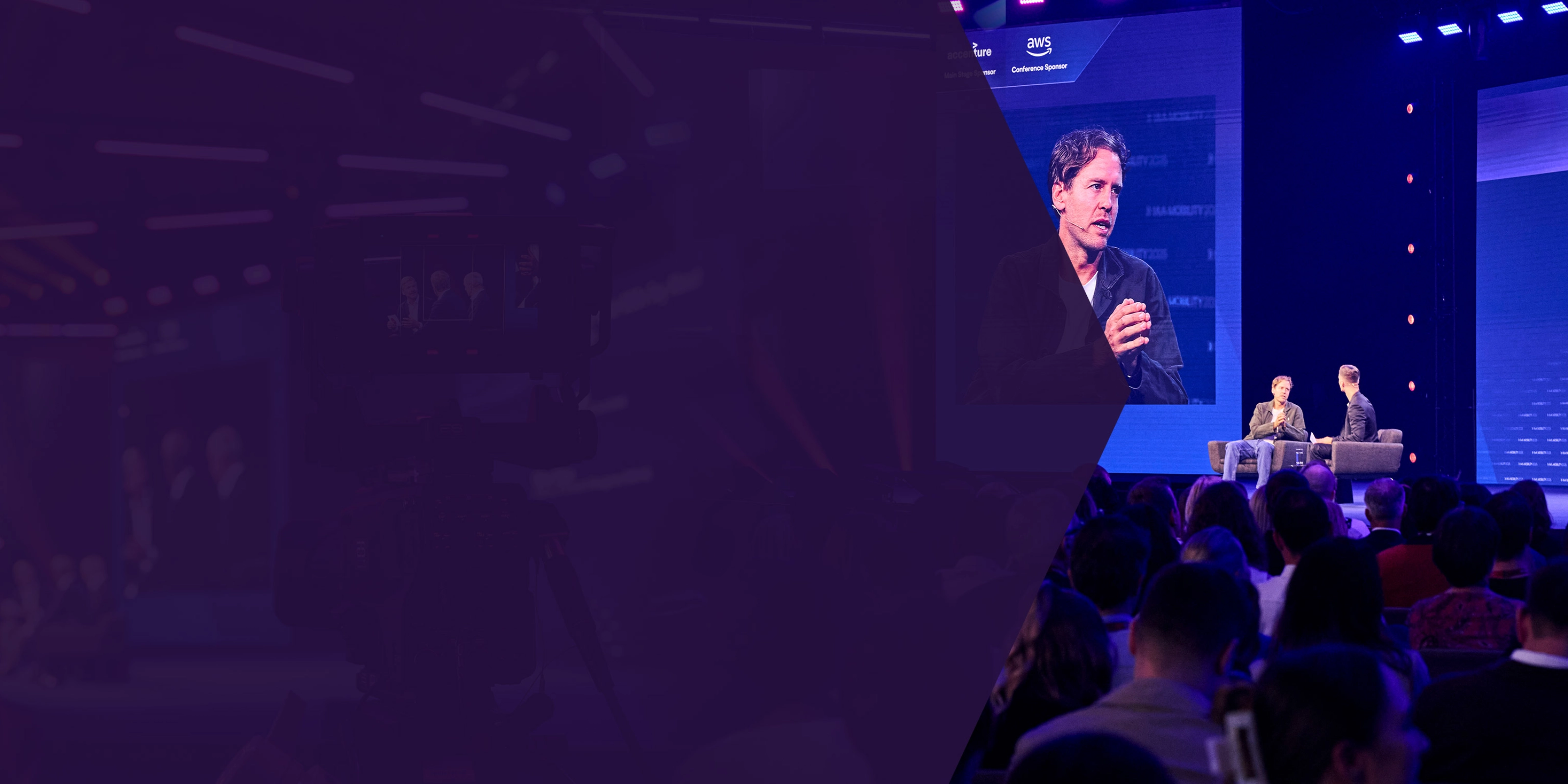 Two men talk on stage during a conference, with one shown on a large screen behind them.
