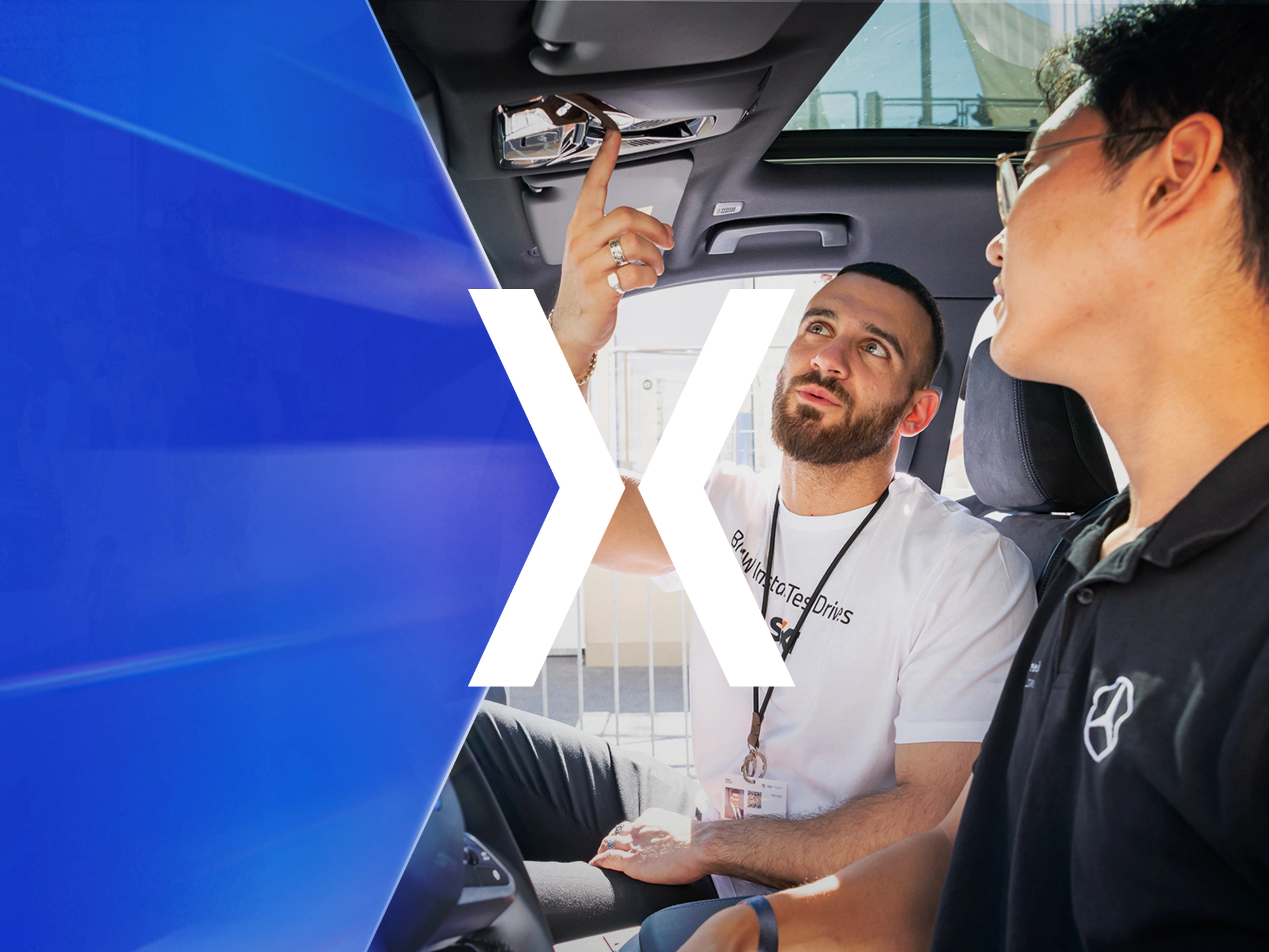 Two men sit in a car while one of them explains a control on the ceiling panel.