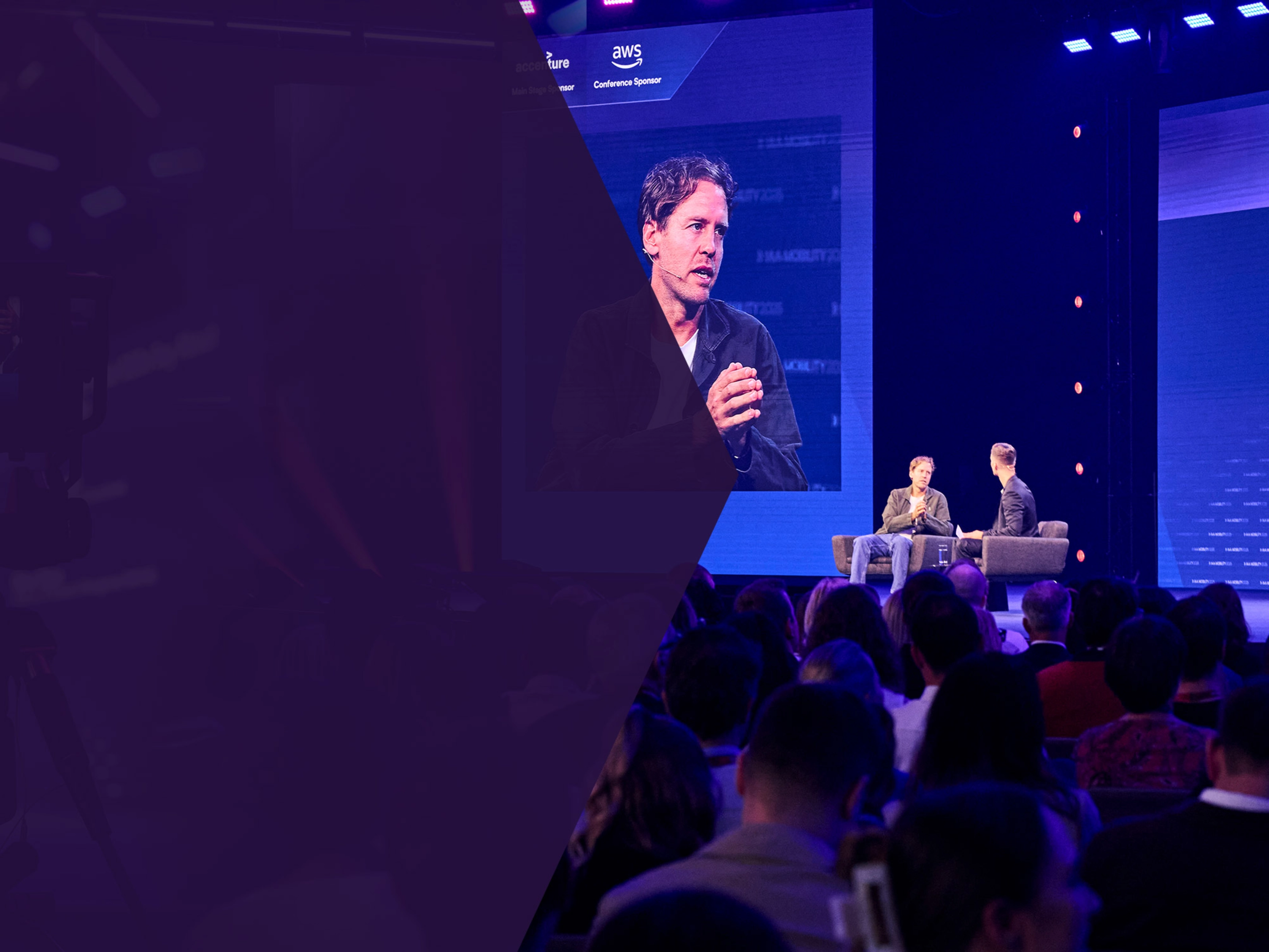 Two men talk on stage during a conference, with one shown on a large screen behind them.