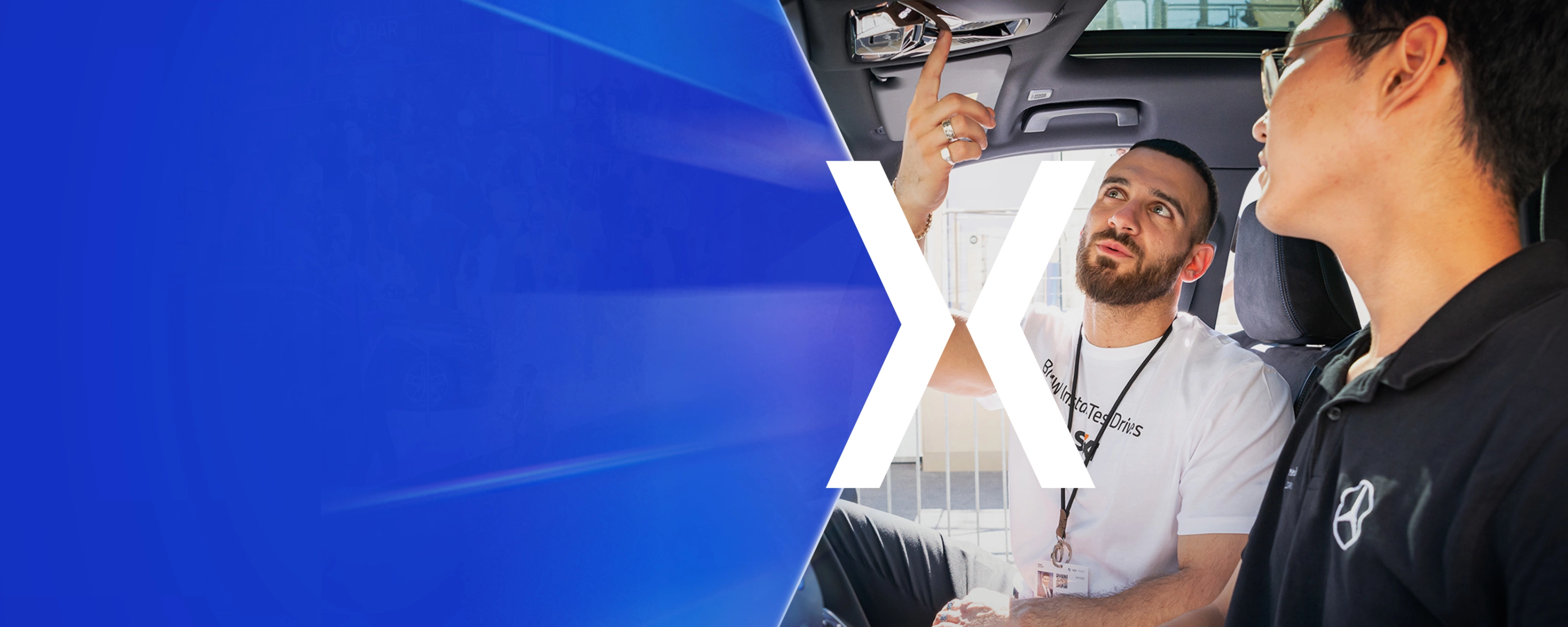 Two men sit in a car while one of them explains a control on the ceiling panel.