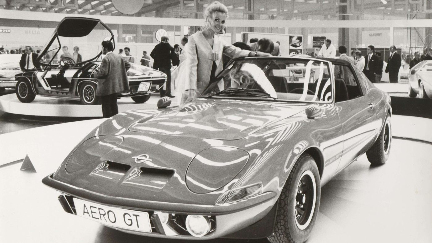 A classic sports car is displayed at an exhibition with a person standing beside it.