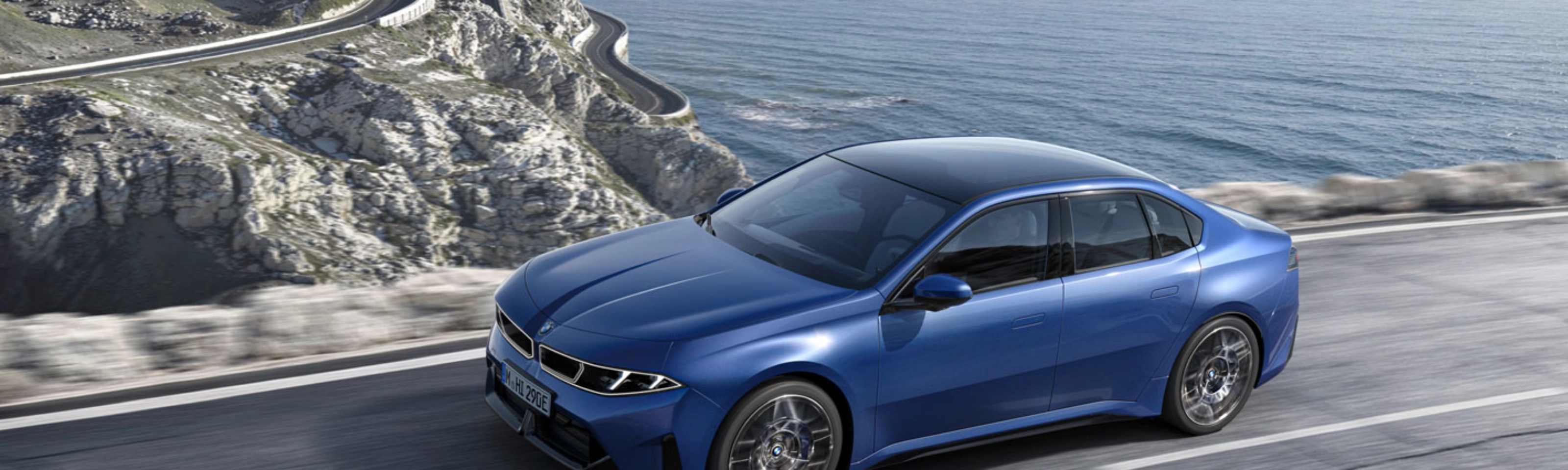 Blue BMW sedan driving along a coastal road with ocean and cliffs in view.