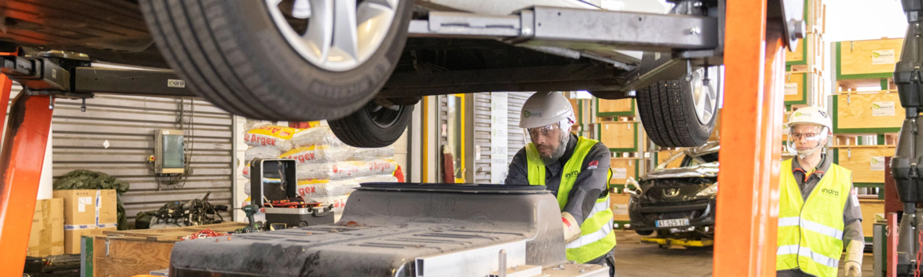 Worker in factory handling EV battery module under vehicle lift.