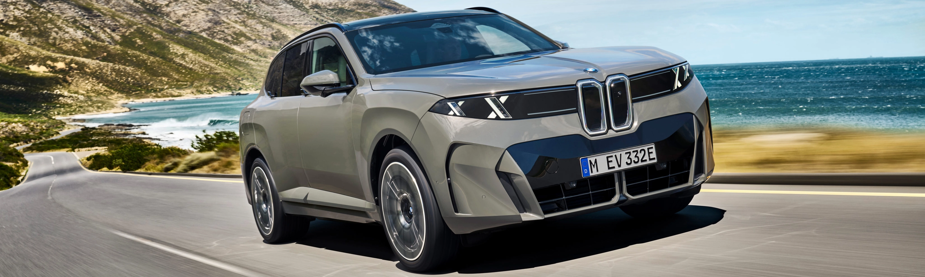 BMW SUV fährt auf einer Küstenstraße mit Bergen und Meer im Hintergrund.