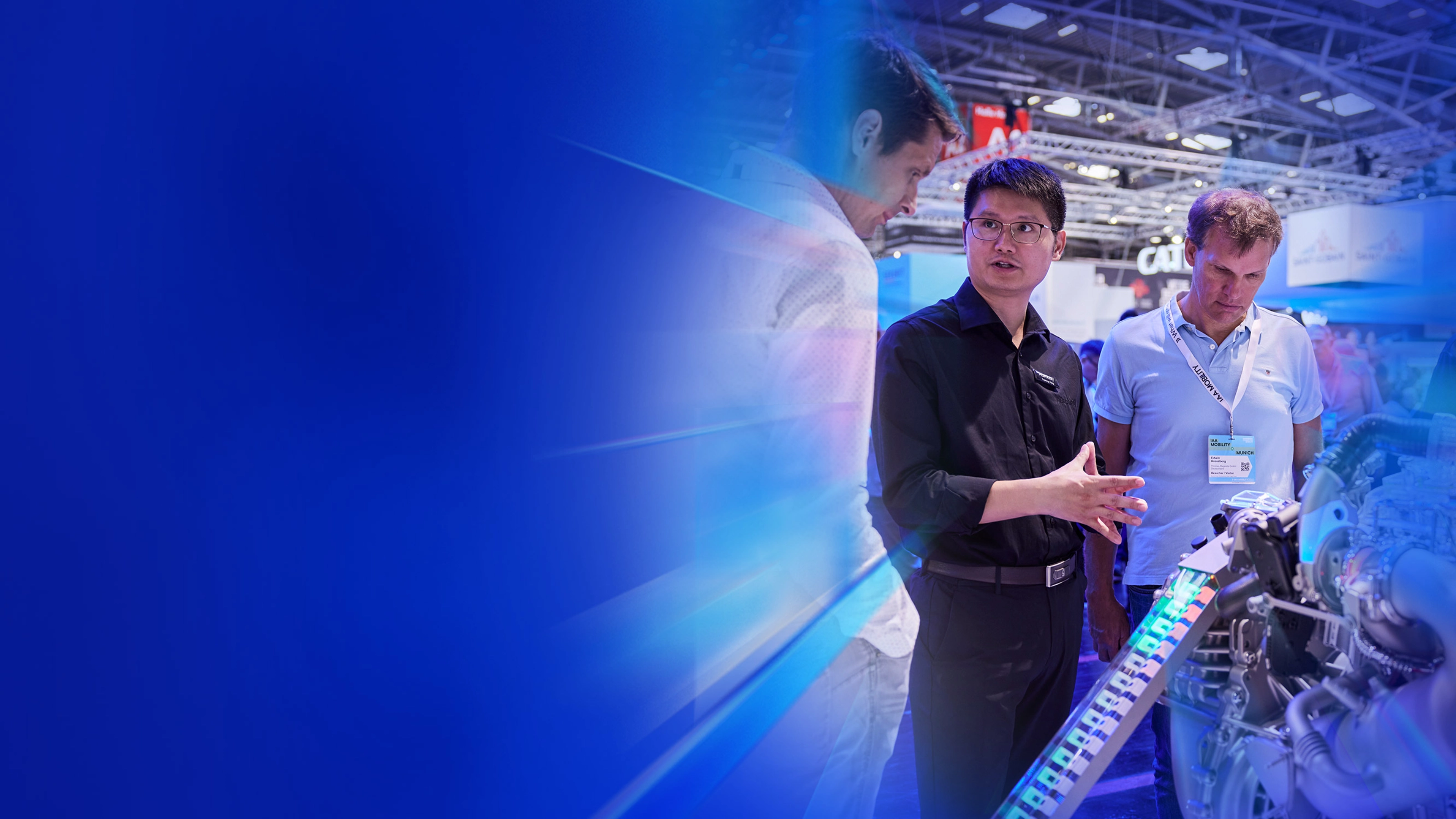 Man explains an engine model to two trade visitors at a technology fair booth.