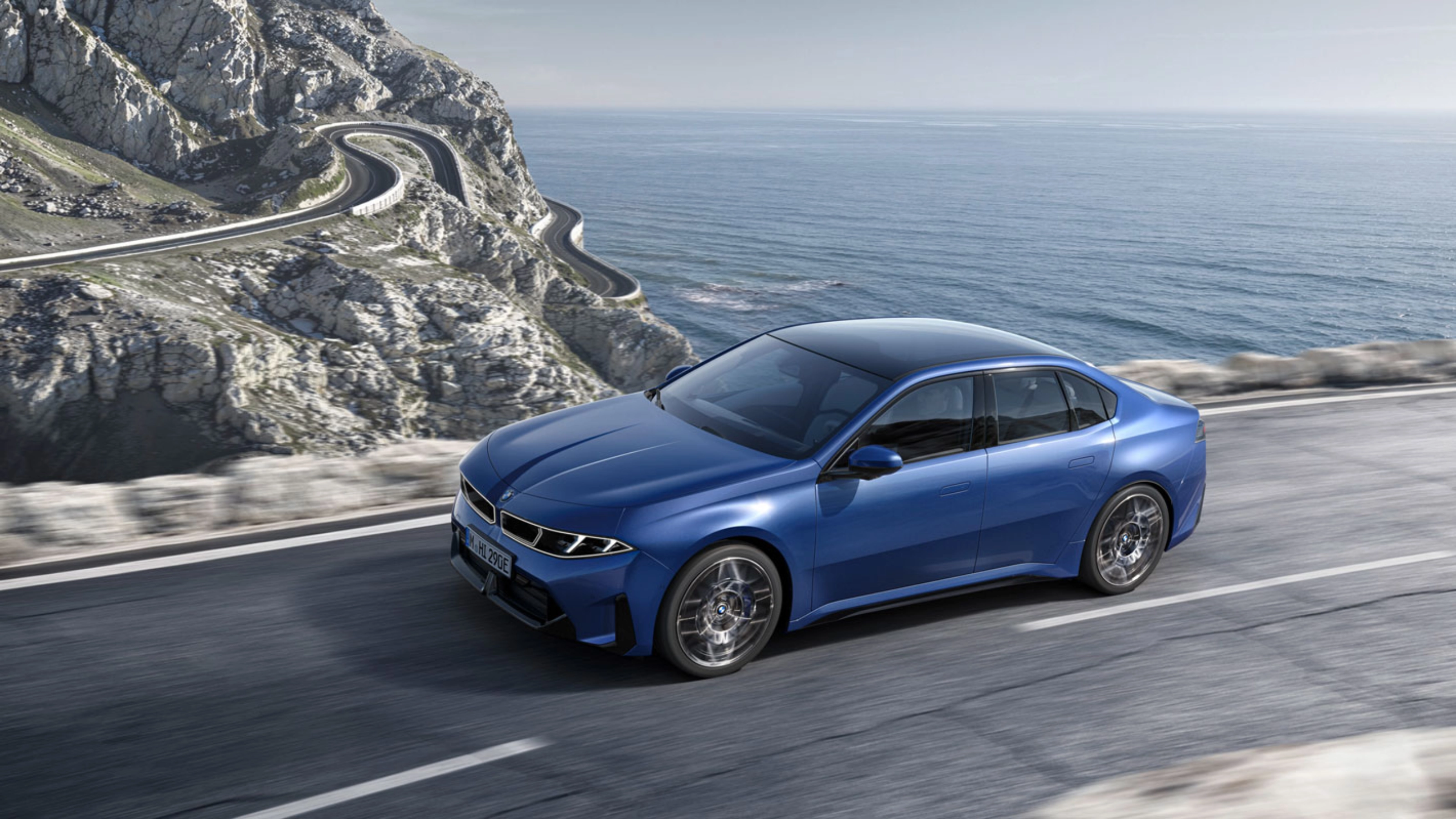 Blue BMW sedan driving along a coastal road with ocean and cliffs in view.