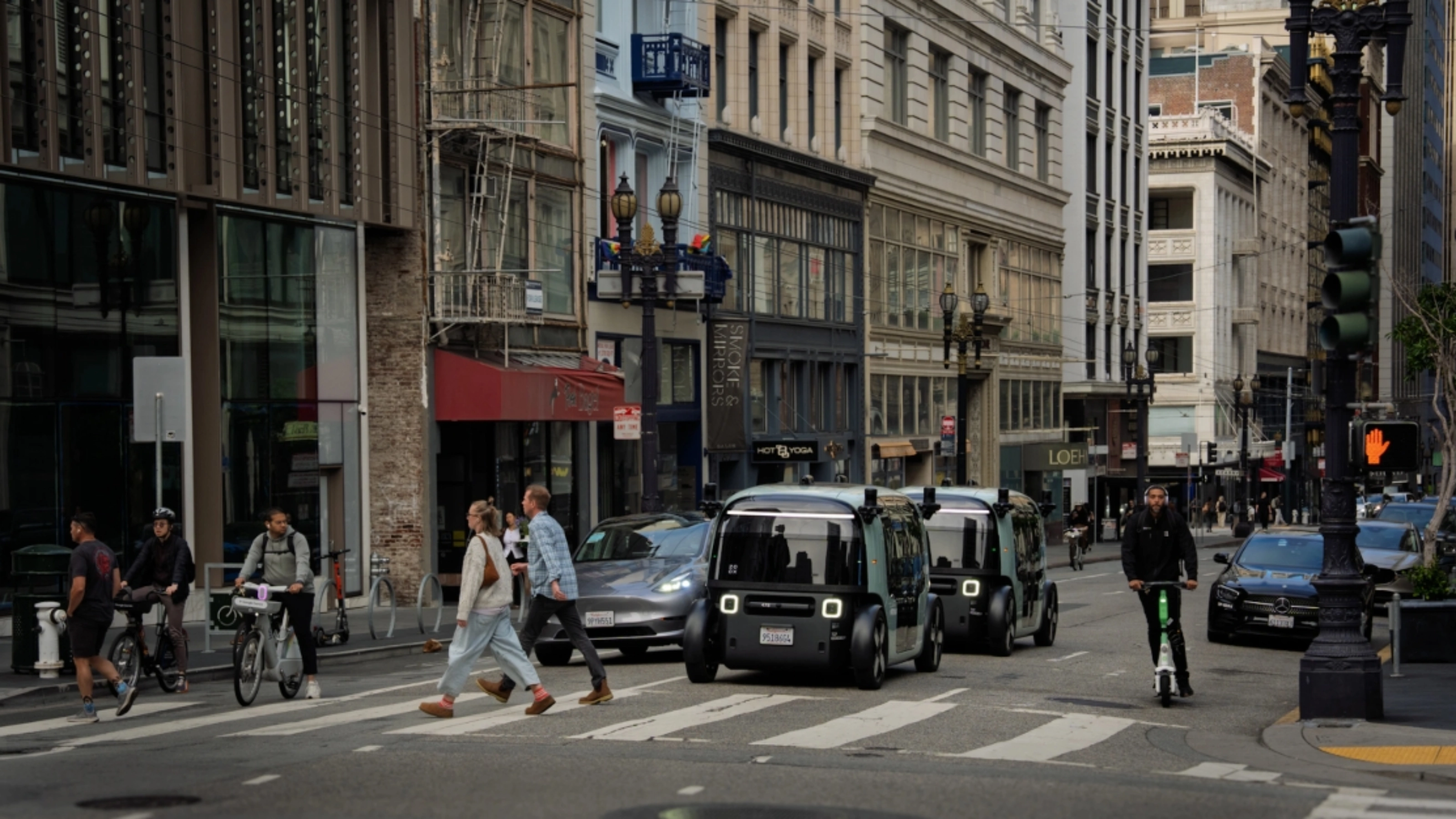 Zoox autonomous shuttle driving through a city intersection with pedestrians nearby.