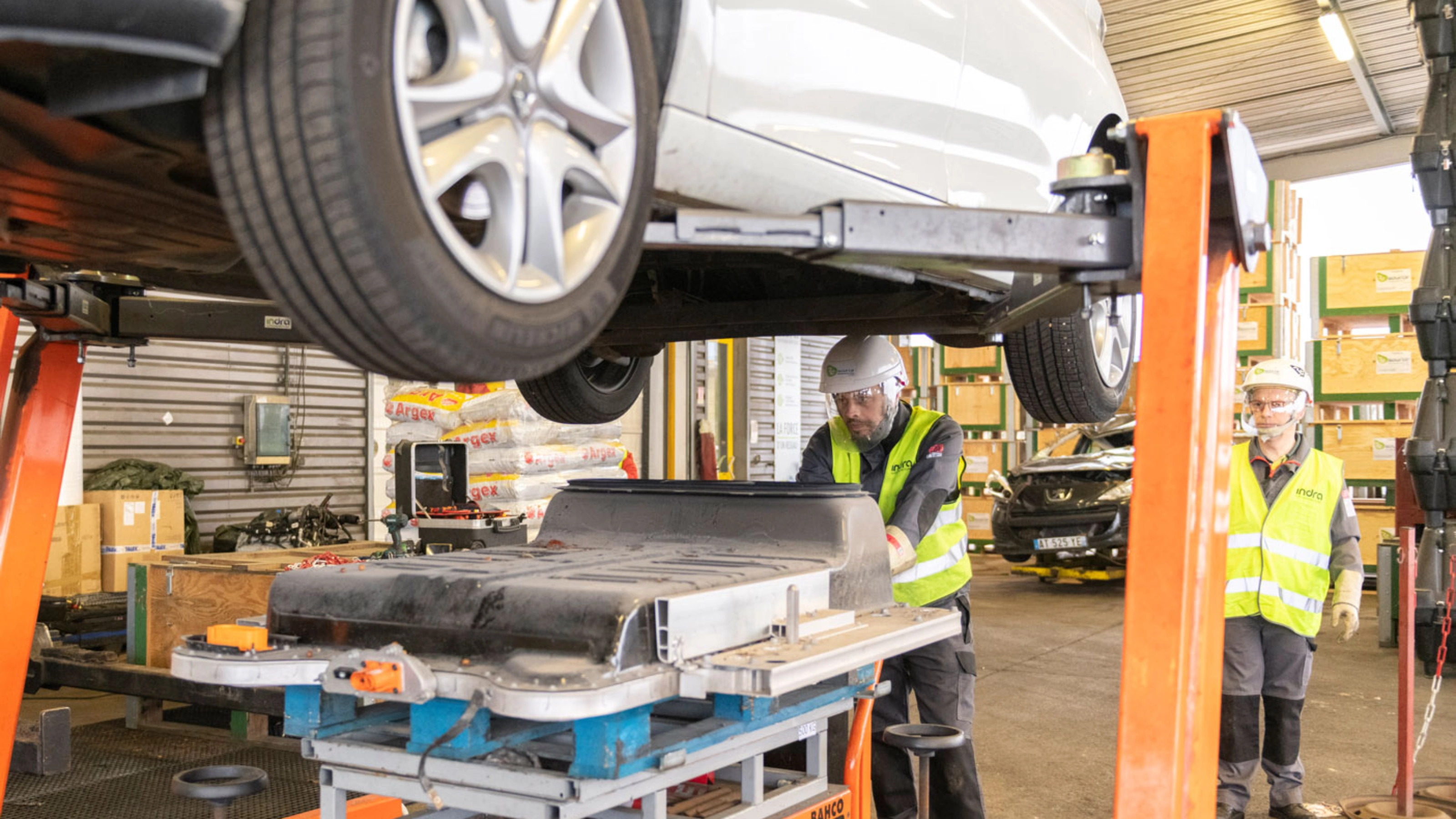 Worker in factory handling EV battery module under vehicle lift.
