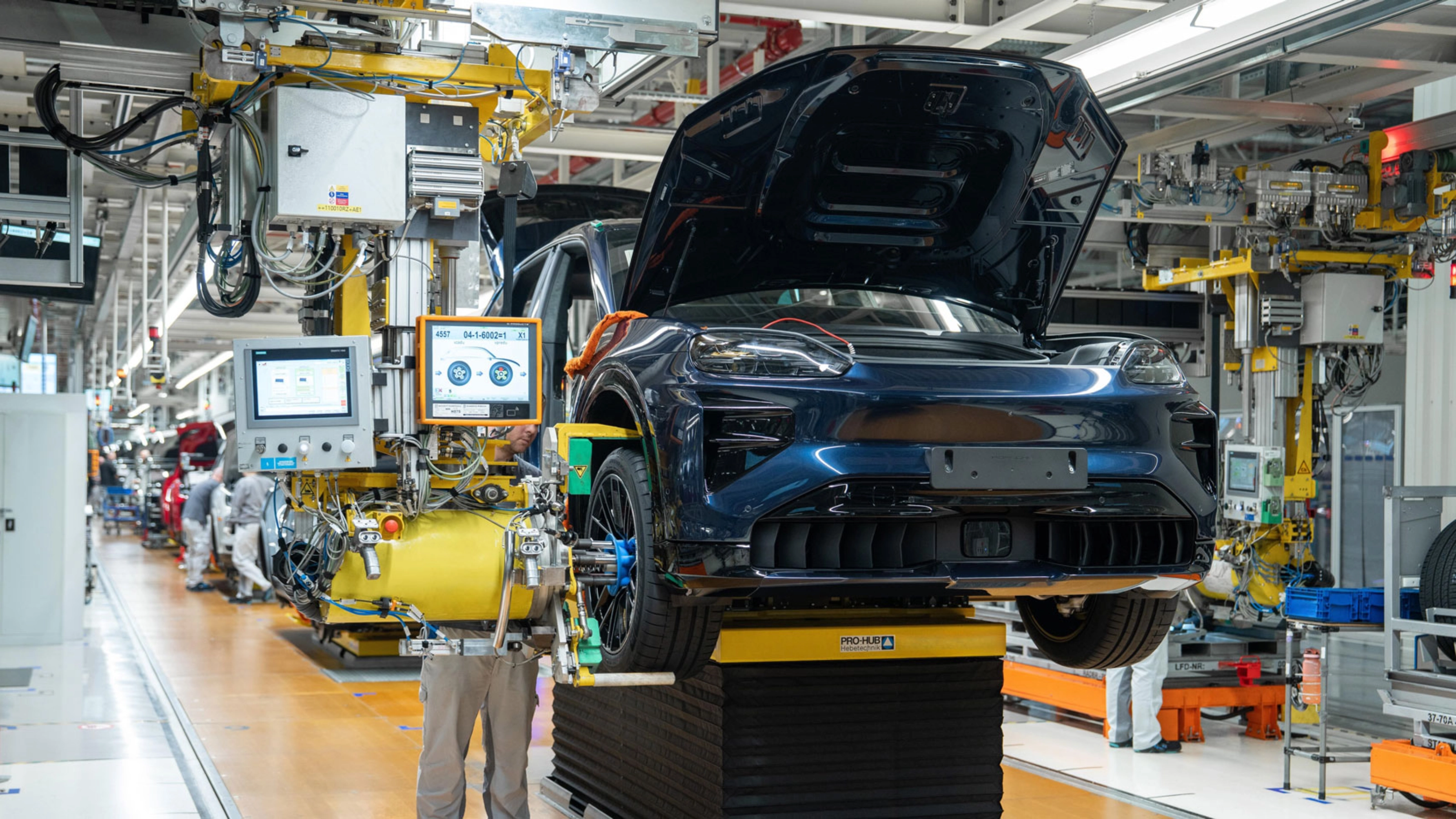 Teilmontiertes Elektroauto auf dem Montageband mit offenem Kofferraum und Maschinen ringsum.