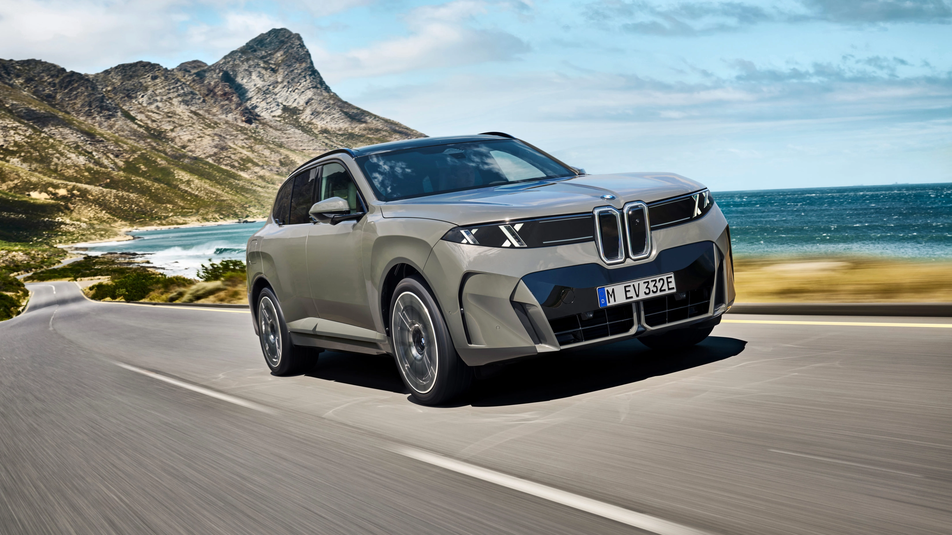 BMW SUV fährt auf einer Küstenstraße mit Bergen und Meer im Hintergrund.