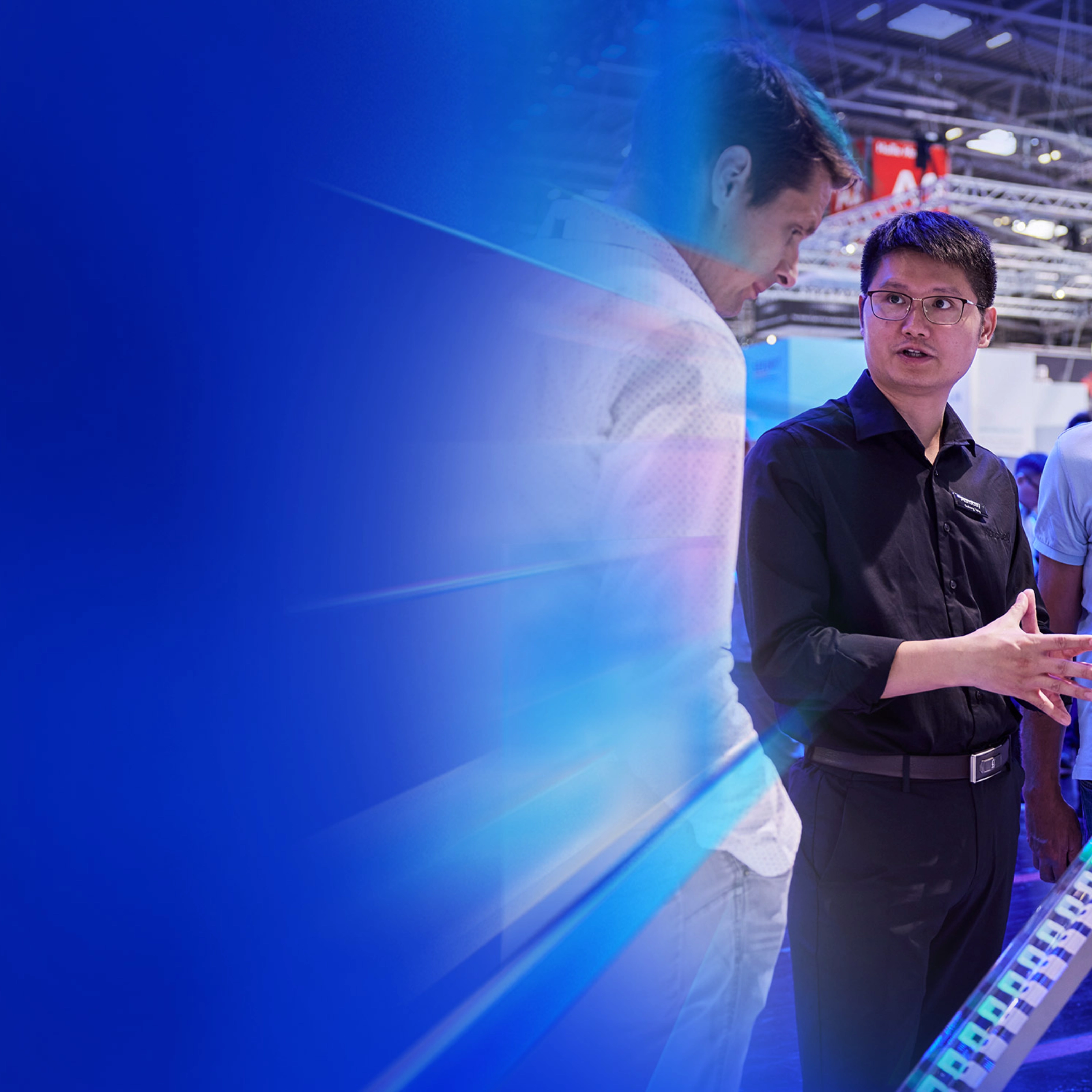 Man explains an engine model to two trade visitors at a technology fair booth.