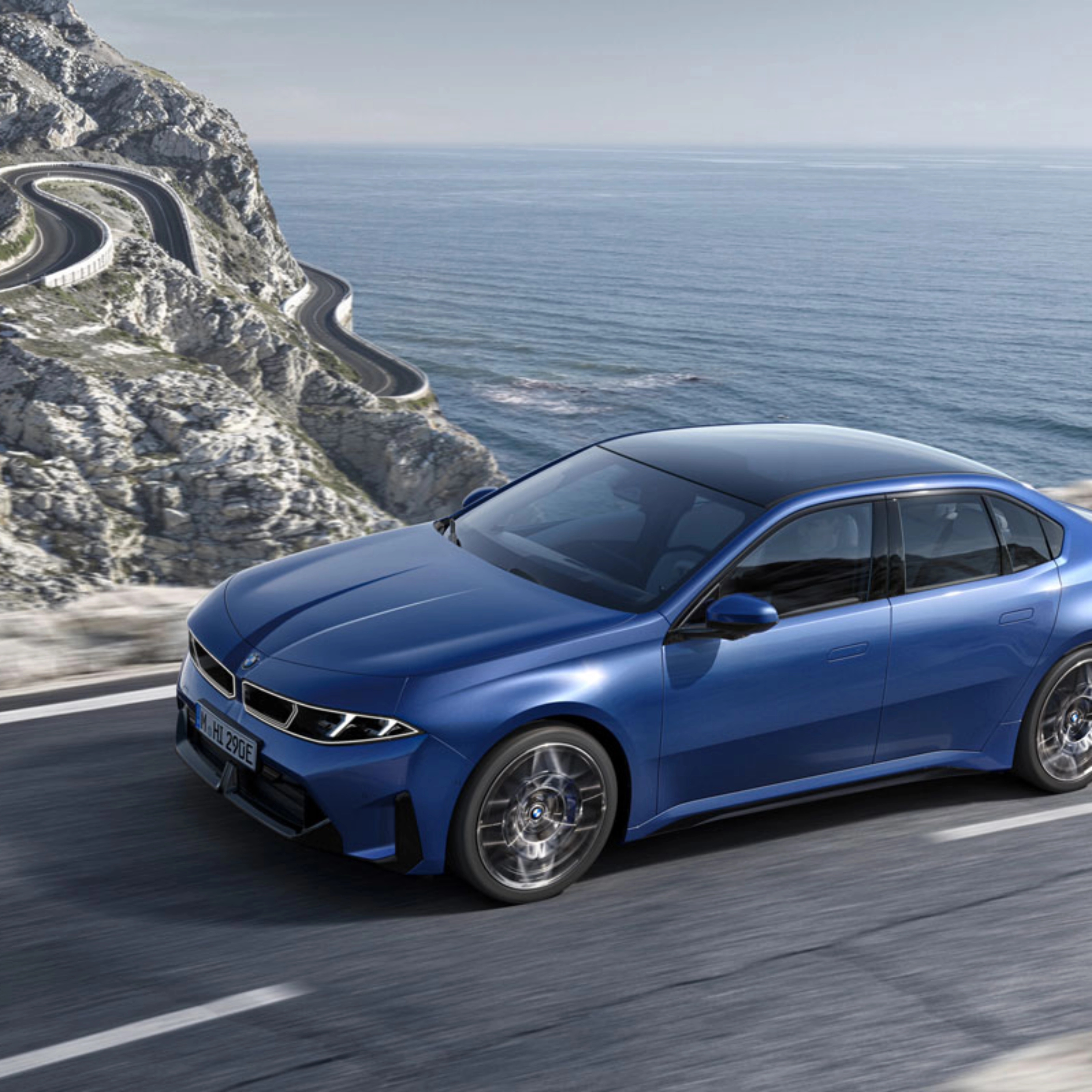 Blue BMW sedan driving along a coastal road with ocean and cliffs in view.
