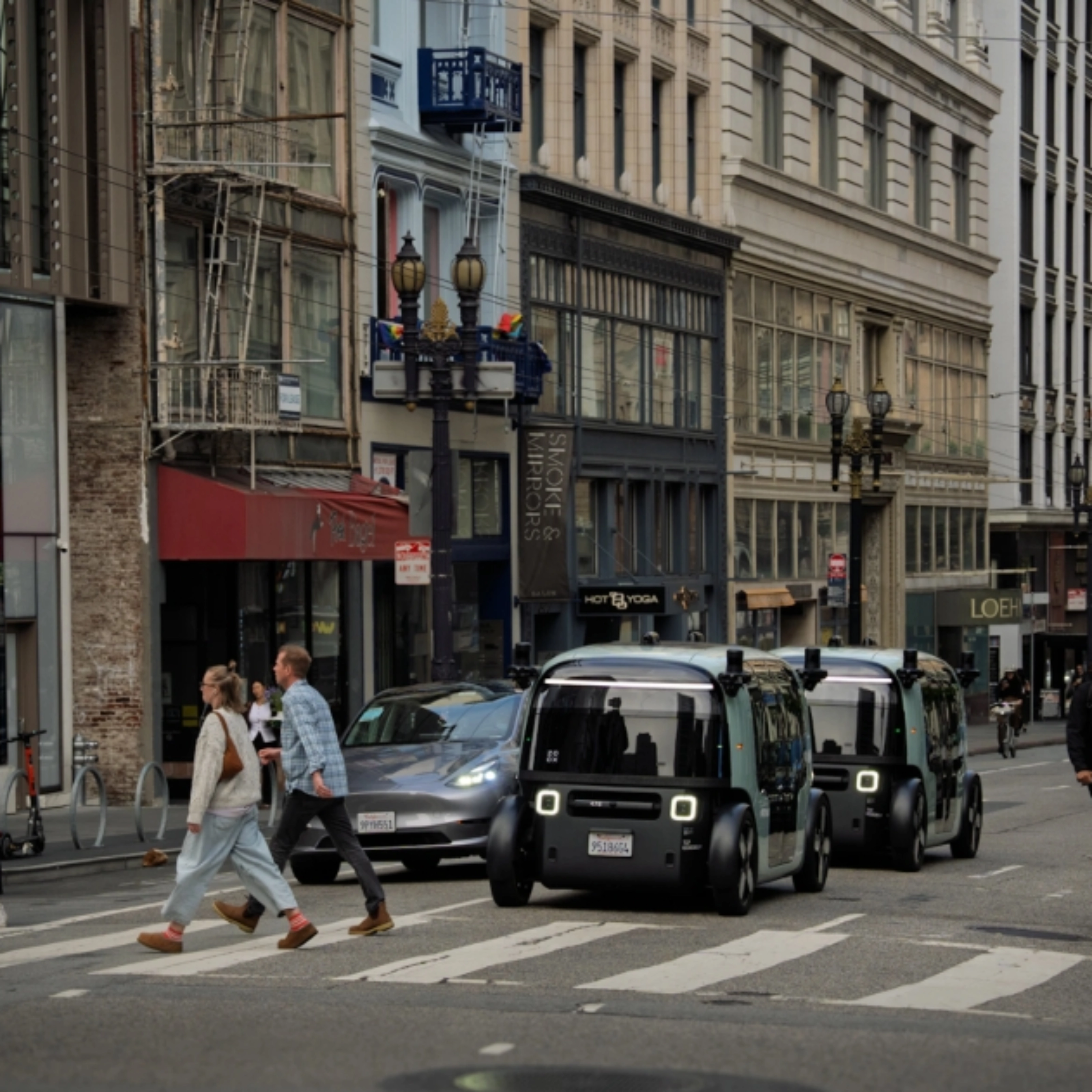 Zoox autonomous shuttle driving through a city intersection with pedestrians nearby.