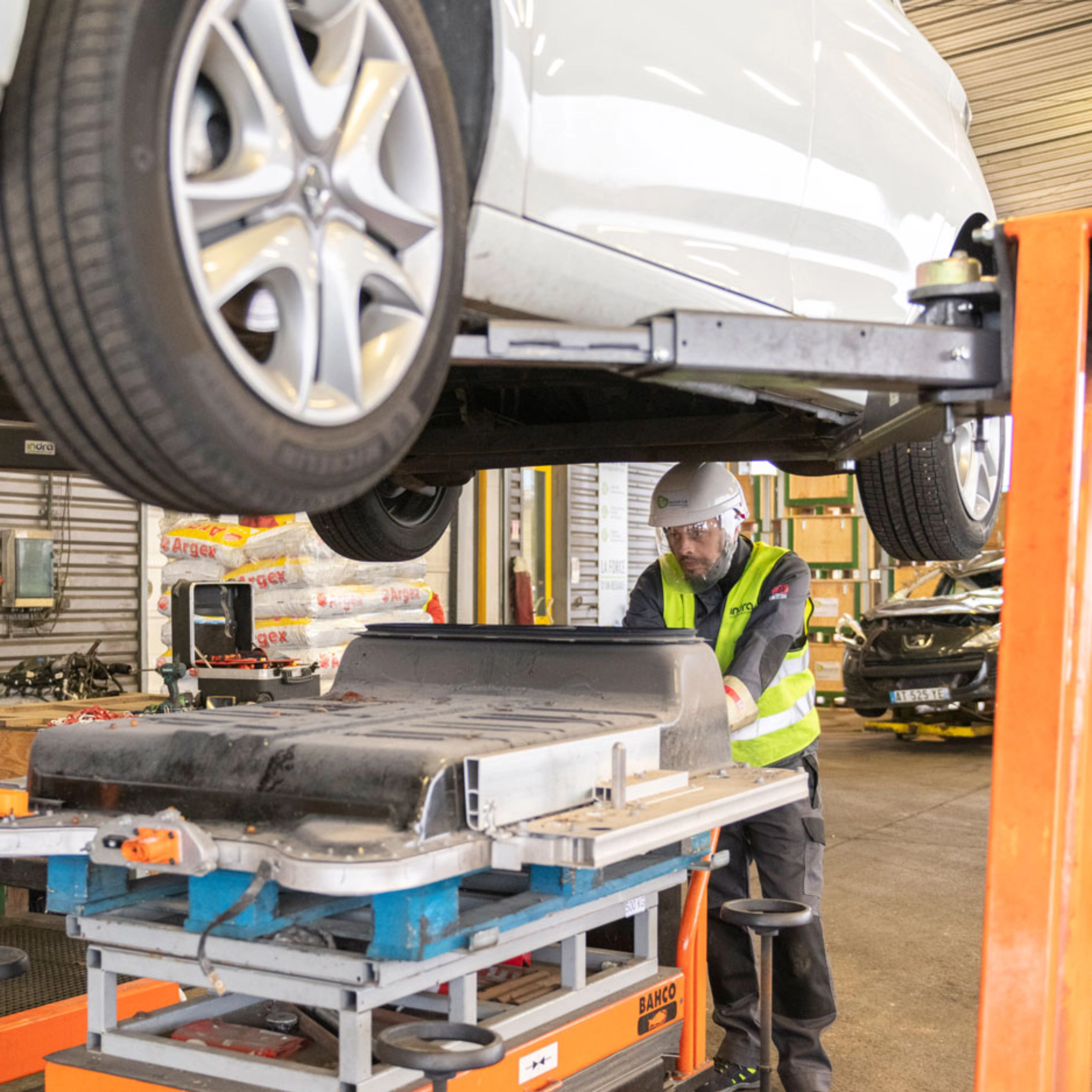 Worker in factory handling EV battery module under vehicle lift.