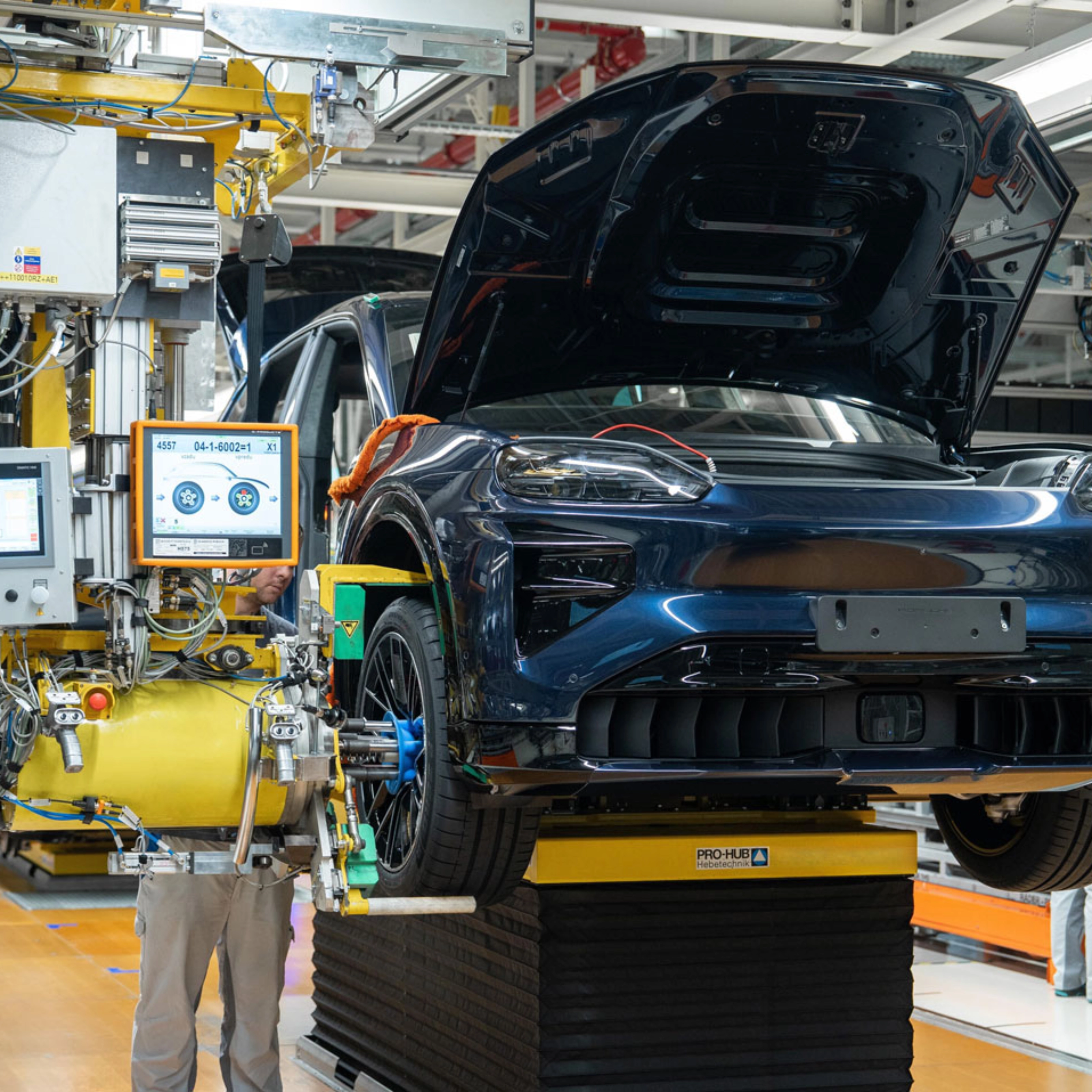 Teilmontiertes Elektroauto auf dem Montageband mit offenem Kofferraum und Maschinen ringsum.