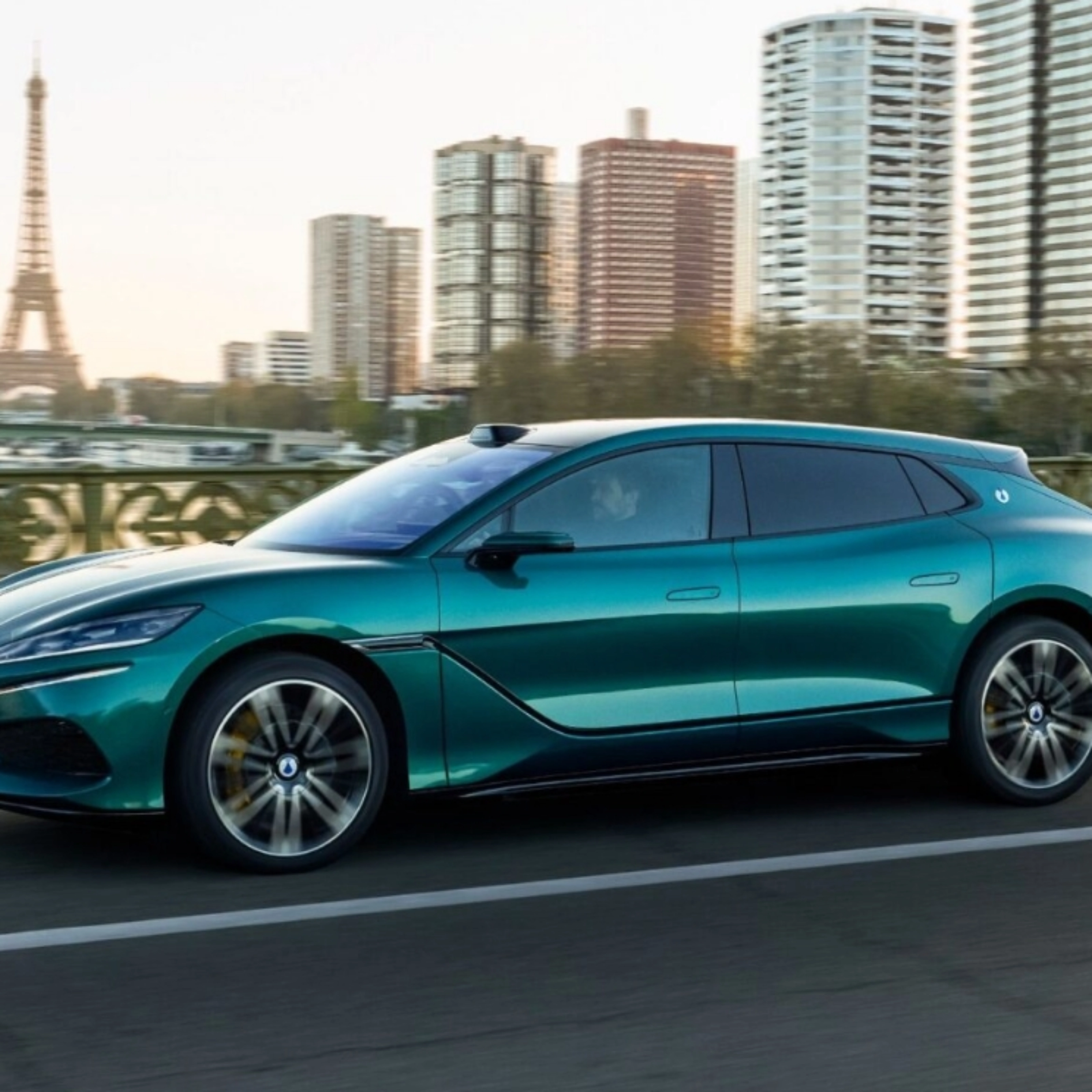 Turquoise BYD electric car driving on a city road with a skyline in the background.