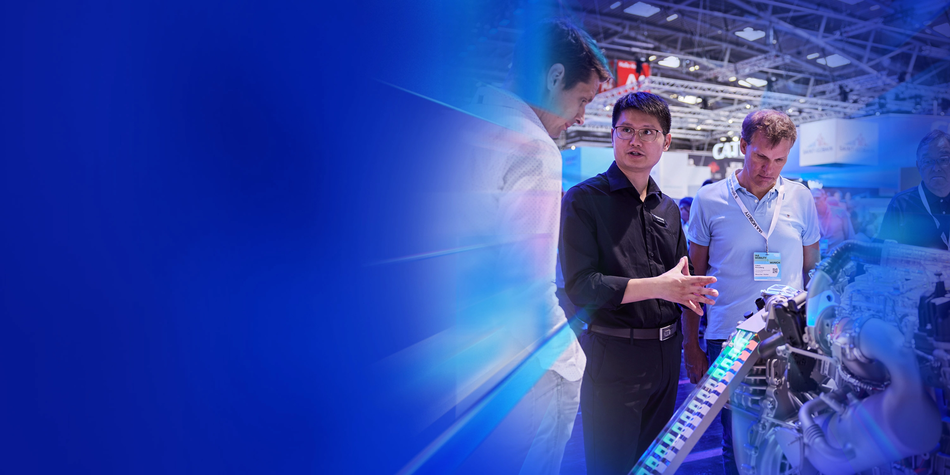 Man explains an engine model to two trade visitors at a technology fair booth.
