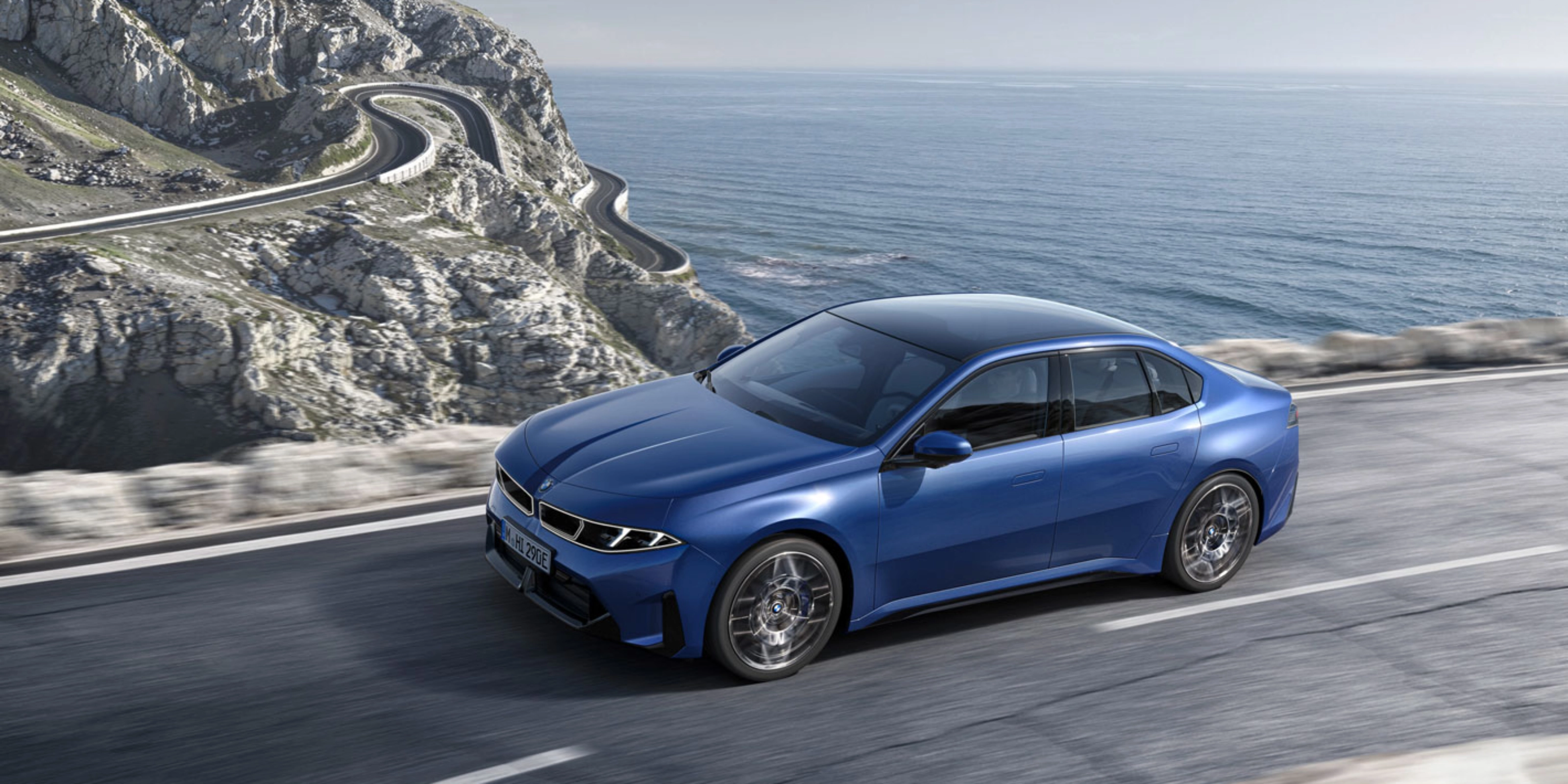 Blue BMW sedan driving along a coastal road with ocean and cliffs in view.