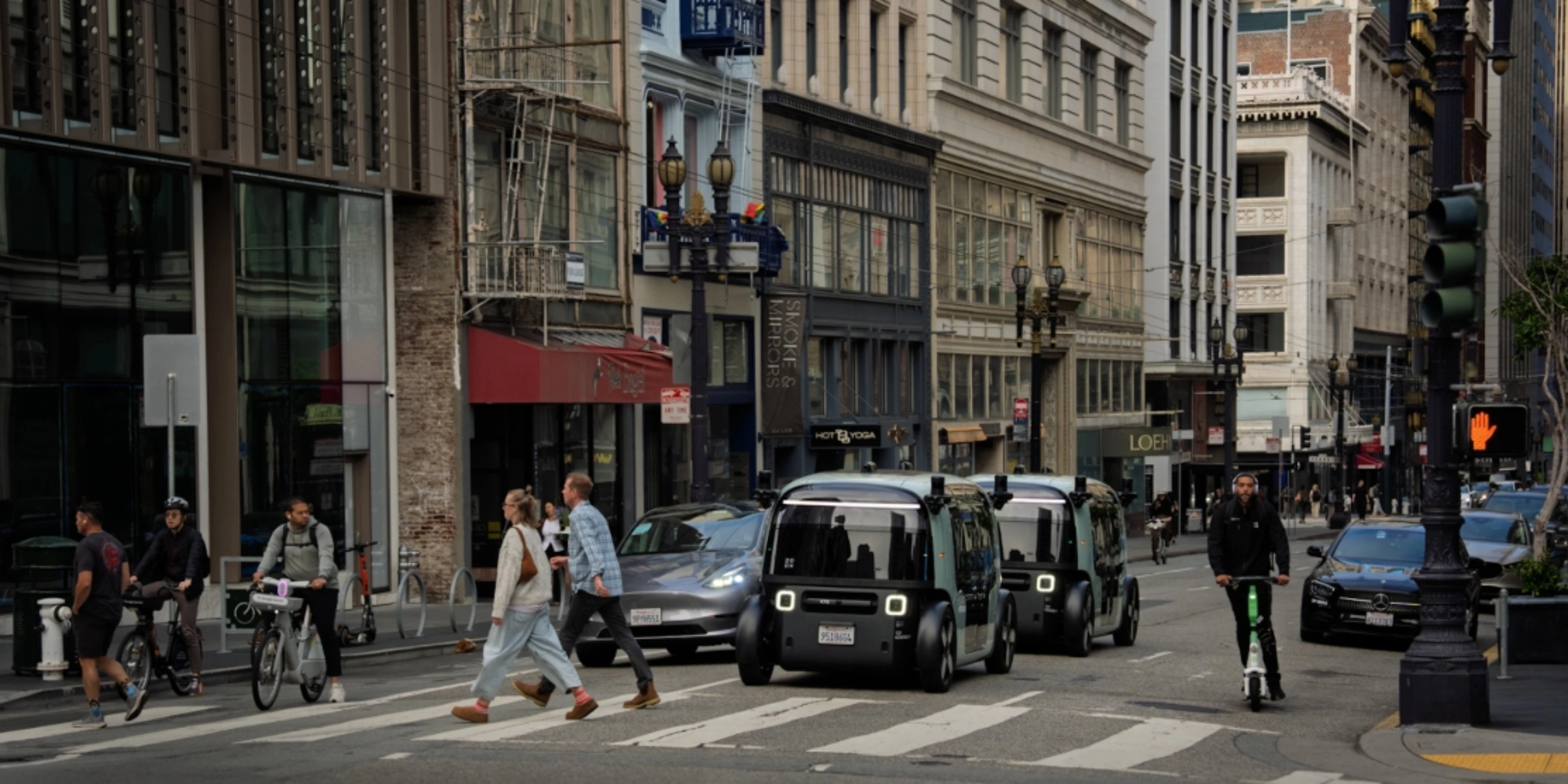 Zoox autonomous shuttle driving through a city intersection with pedestrians nearby.