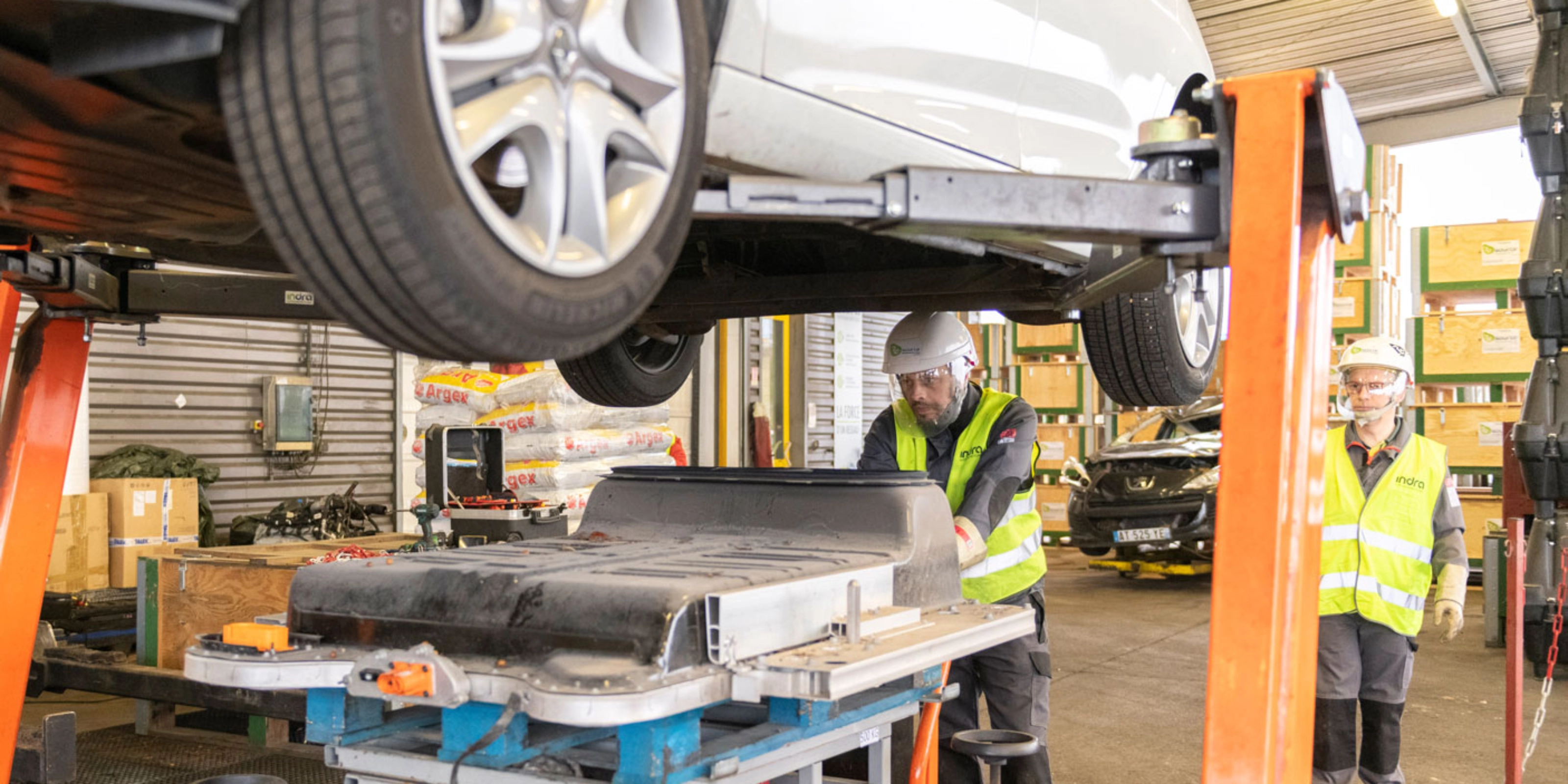 Worker in factory handling EV battery module under vehicle lift.