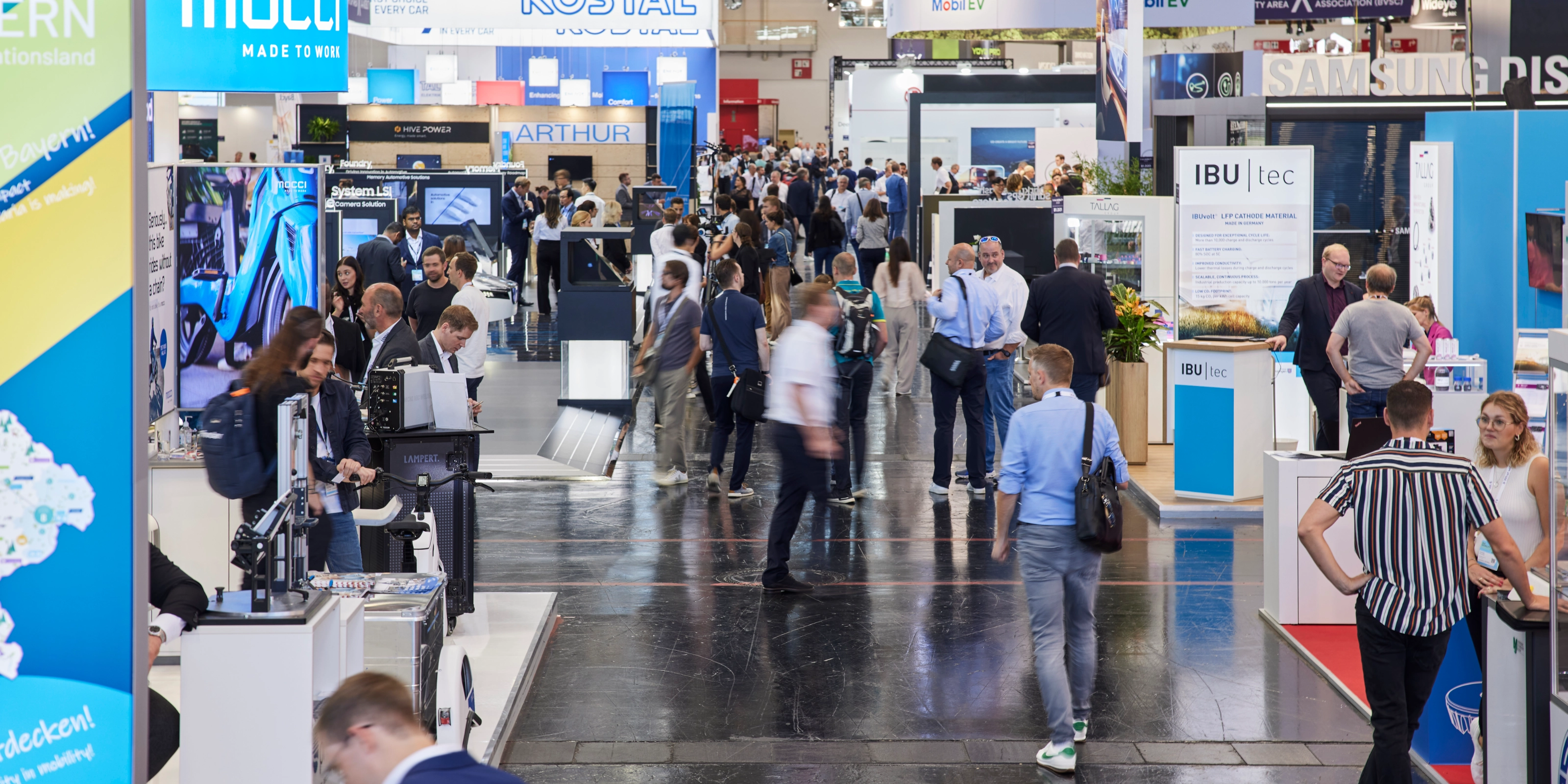 Busy trade fair hall with numerous booths, digital displays, and visitors walking around.