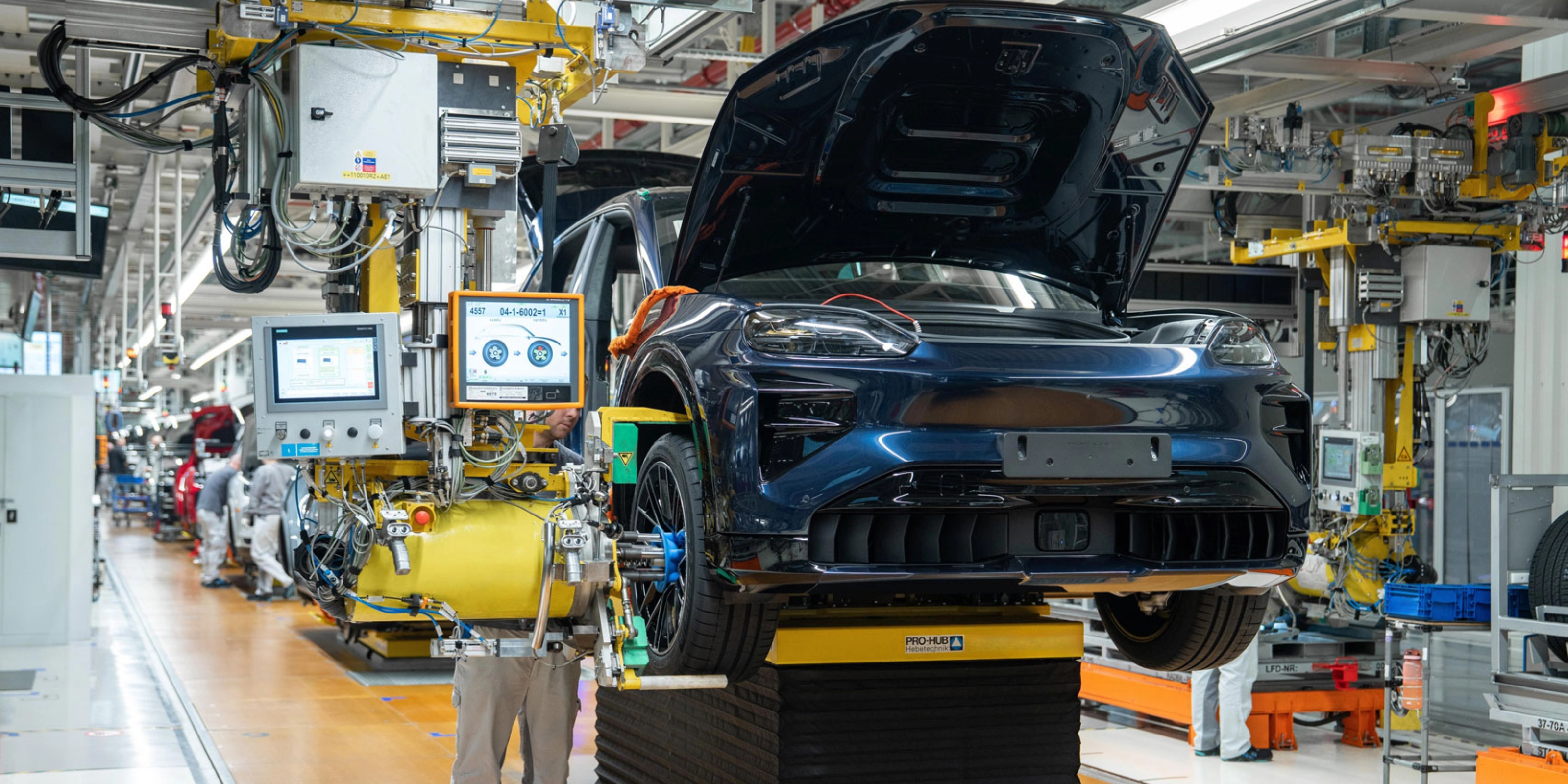 Teilmontiertes Elektroauto auf dem Montageband mit offenem Kofferraum und Maschinen ringsum.