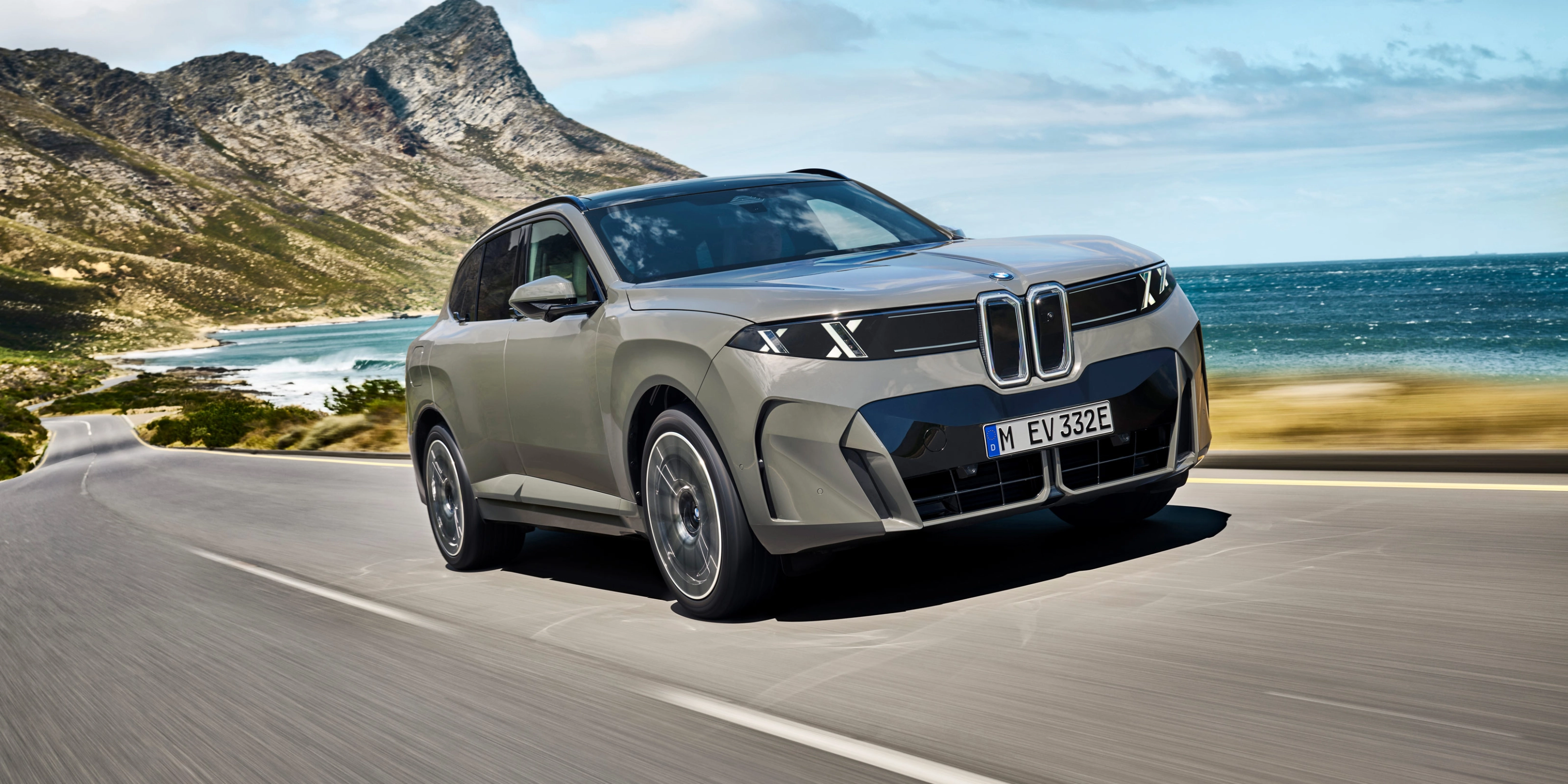 BMW SUV fährt auf einer Küstenstraße mit Bergen und Meer im Hintergrund.