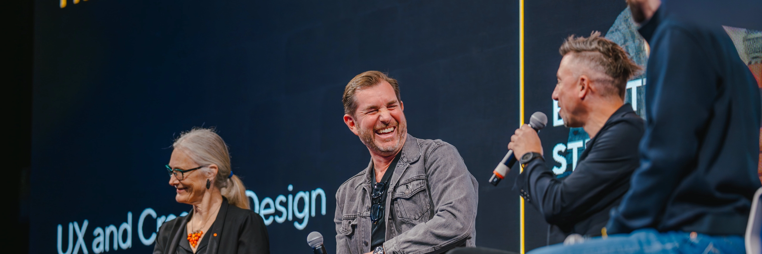 Three people with microphones laugh during a panel discussion on stage.