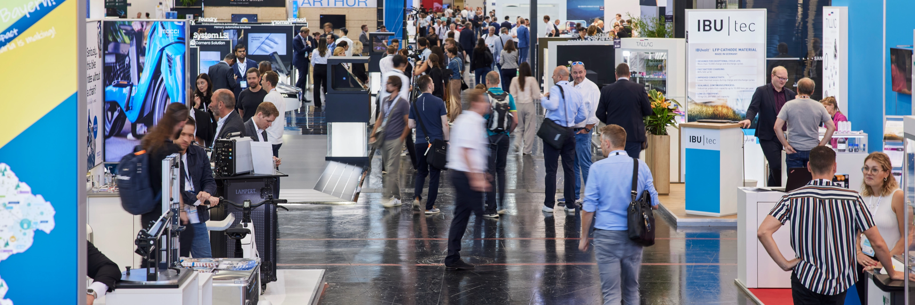 Busy trade fair hall with numerous booths, digital displays, and visitors walking around.