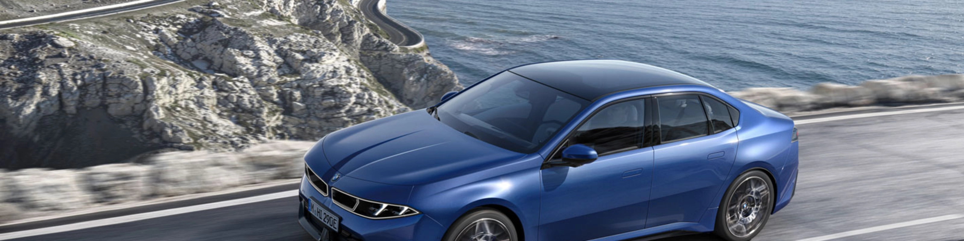 Blue BMW sedan driving along a coastal road with ocean and cliffs in view.