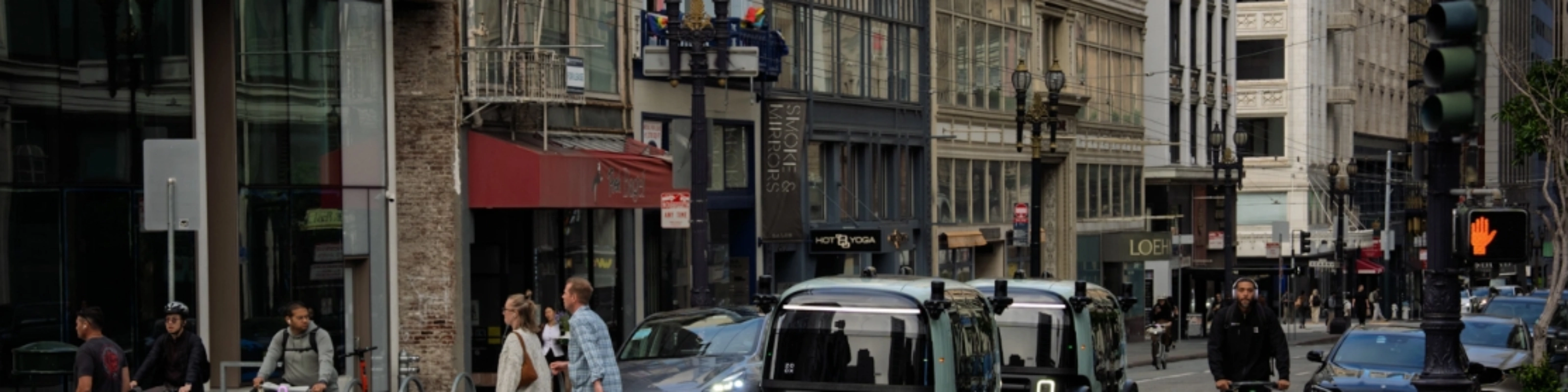 Zoox autonomous shuttle driving through a city intersection with pedestrians nearby.