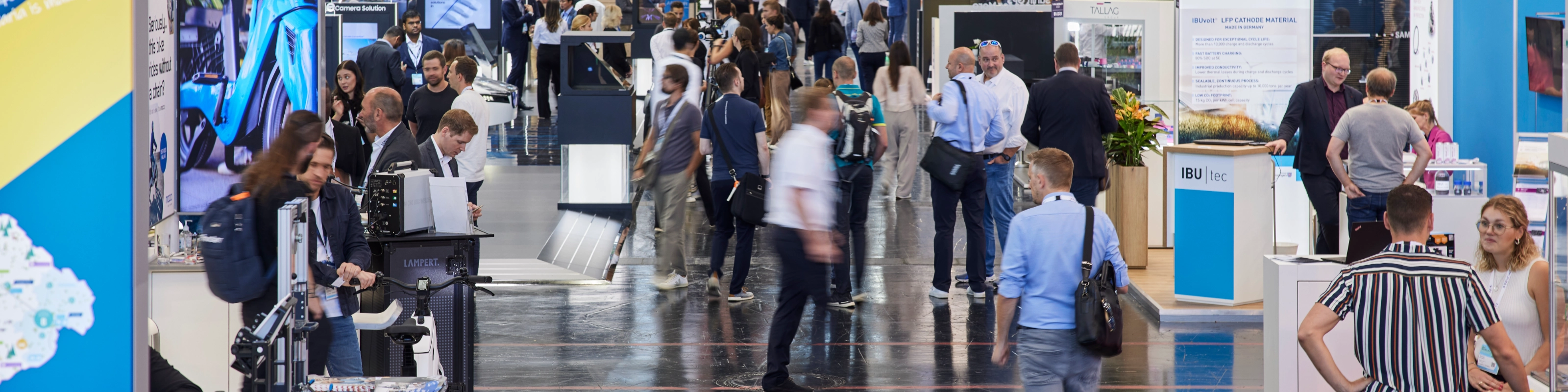 Busy trade fair hall with numerous booths, digital displays, and visitors walking around.