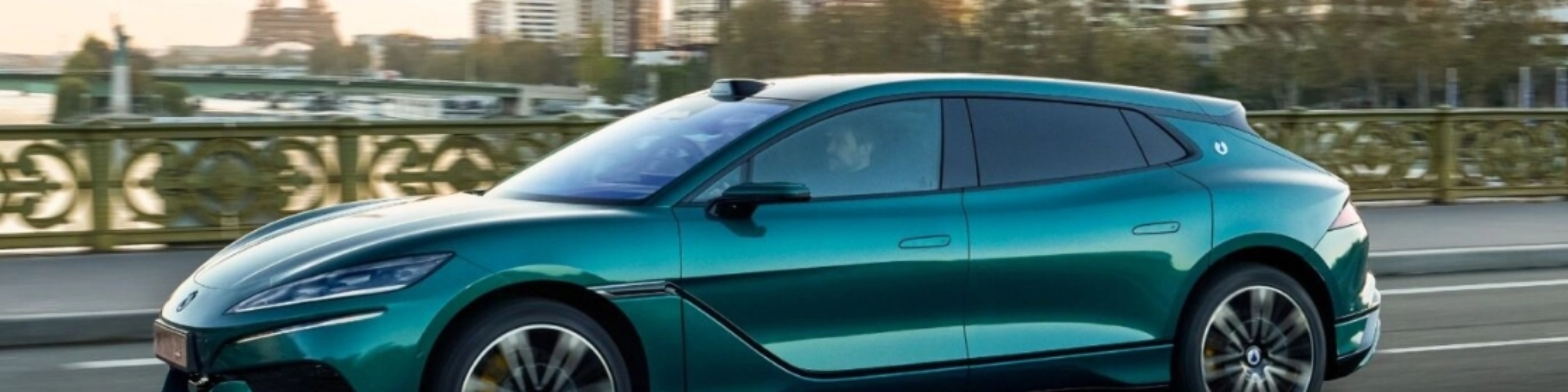 Turquoise BYD electric car driving on a city road with a skyline in the background.