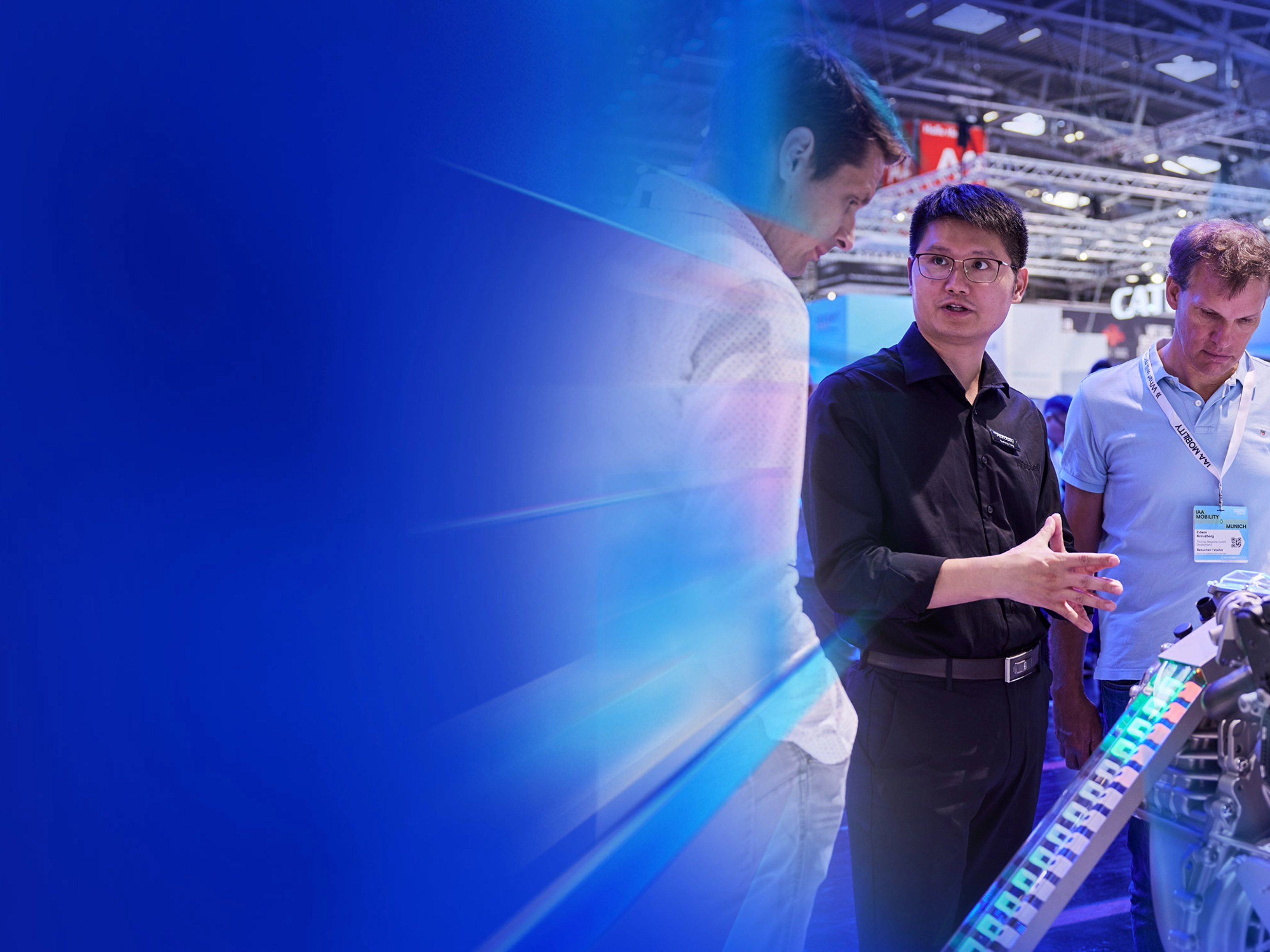 Man explains an engine model to two trade visitors at a technology fair booth.