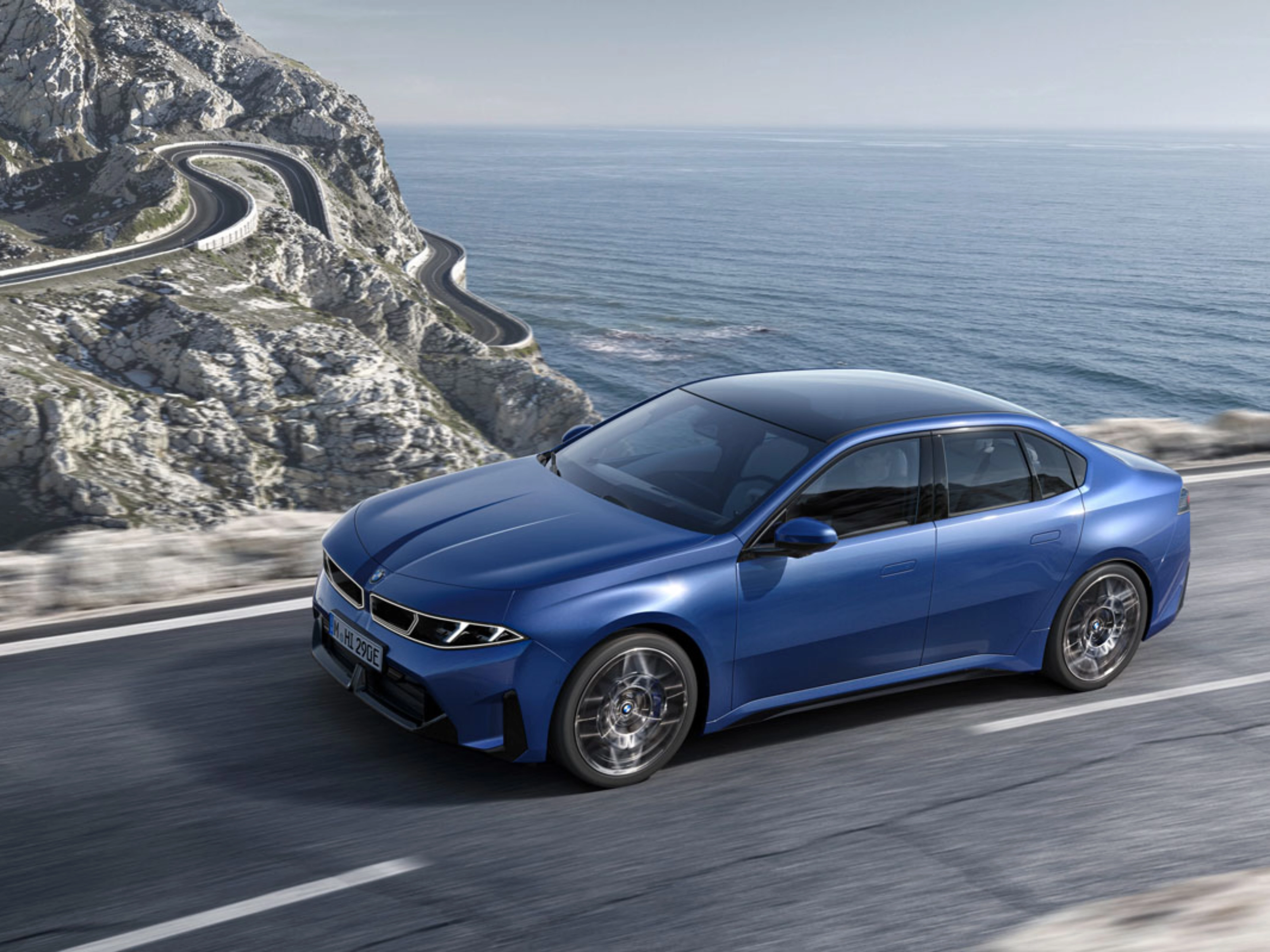 Blue BMW sedan driving along a coastal road with ocean and cliffs in view.