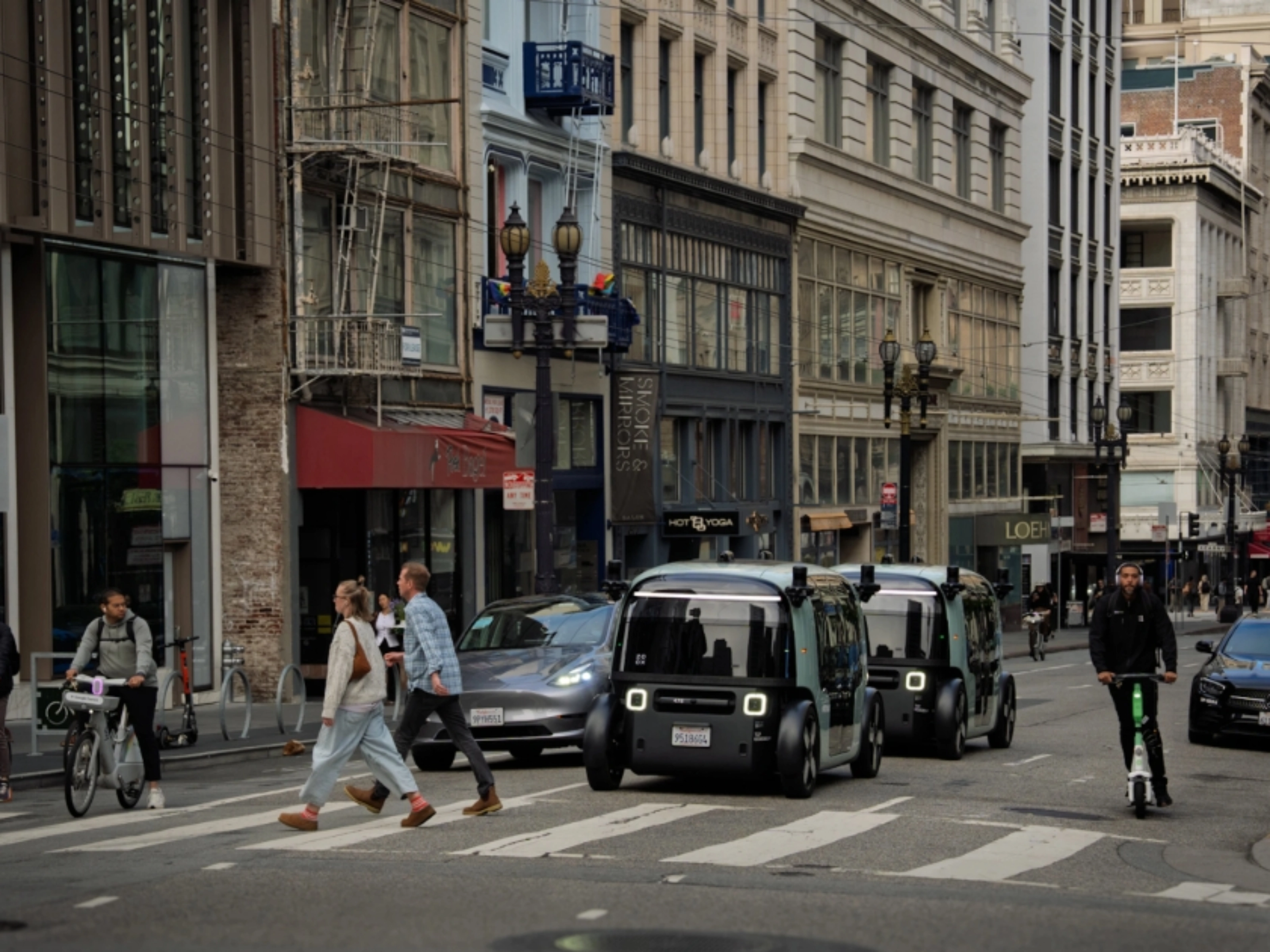 Zoox autonomous shuttle driving through a city intersection with pedestrians nearby.