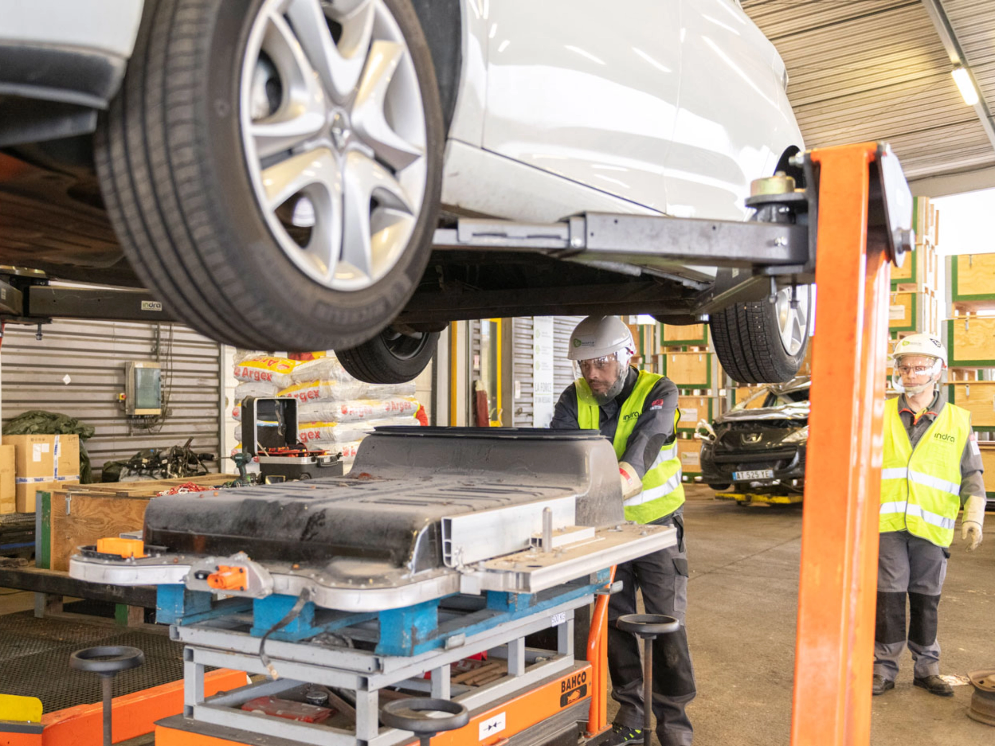 Worker in factory handling EV battery module under vehicle lift.