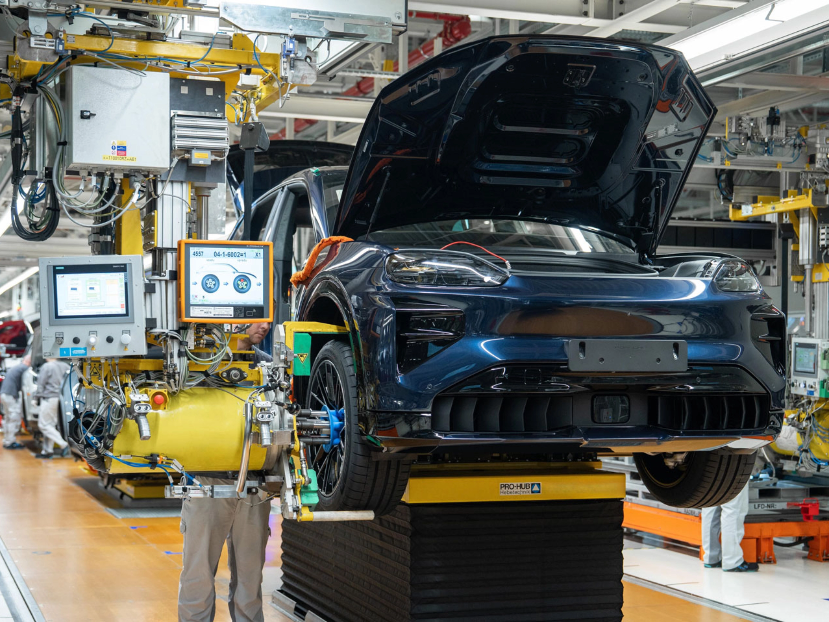 Teilmontiertes Elektroauto auf dem Montageband mit offenem Kofferraum und Maschinen ringsum.