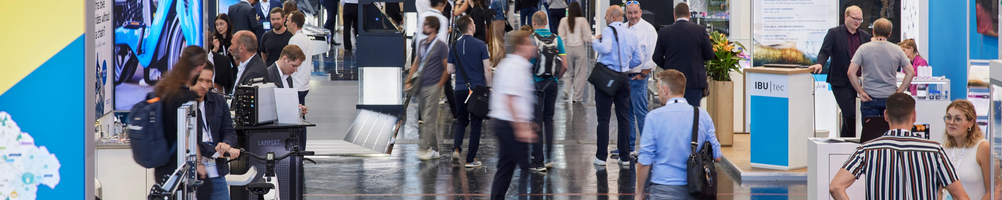 Busy trade fair hall with numerous booths, digital displays, and visitors walking around.