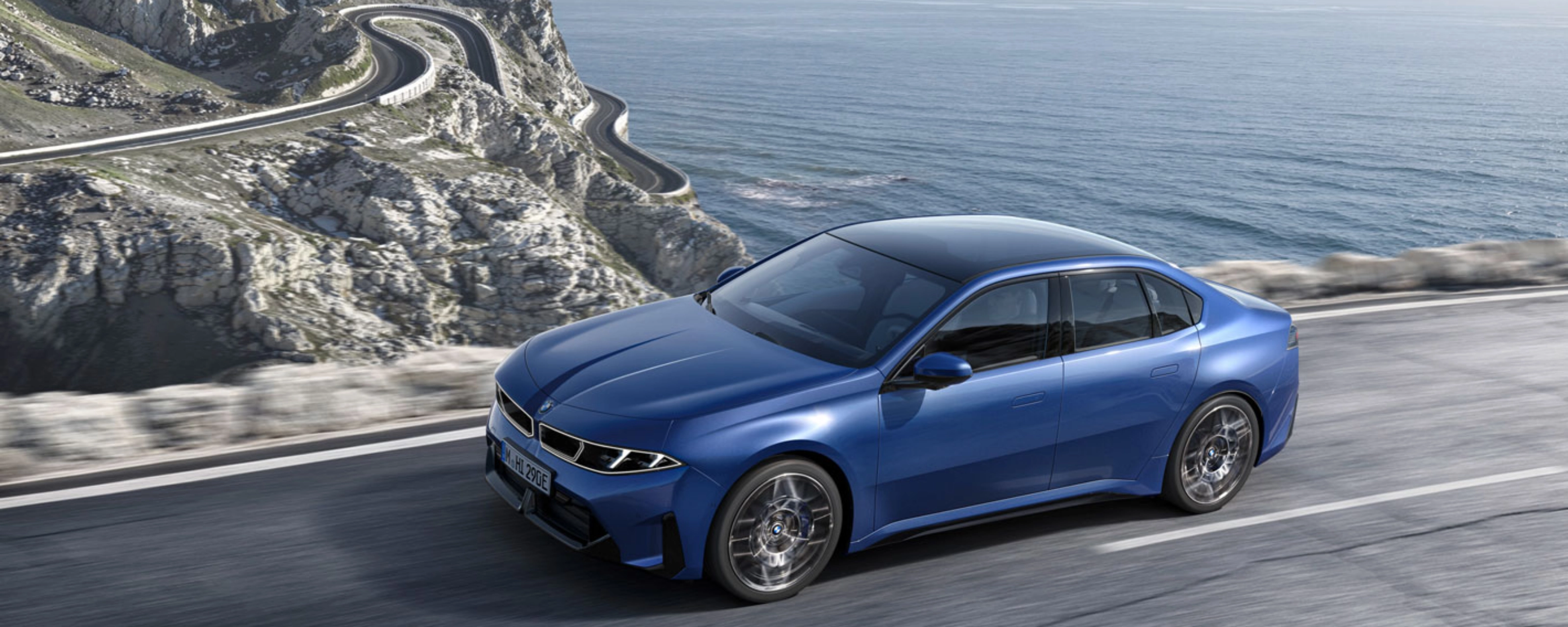 Blue BMW sedan driving along a coastal road with ocean and cliffs in view.