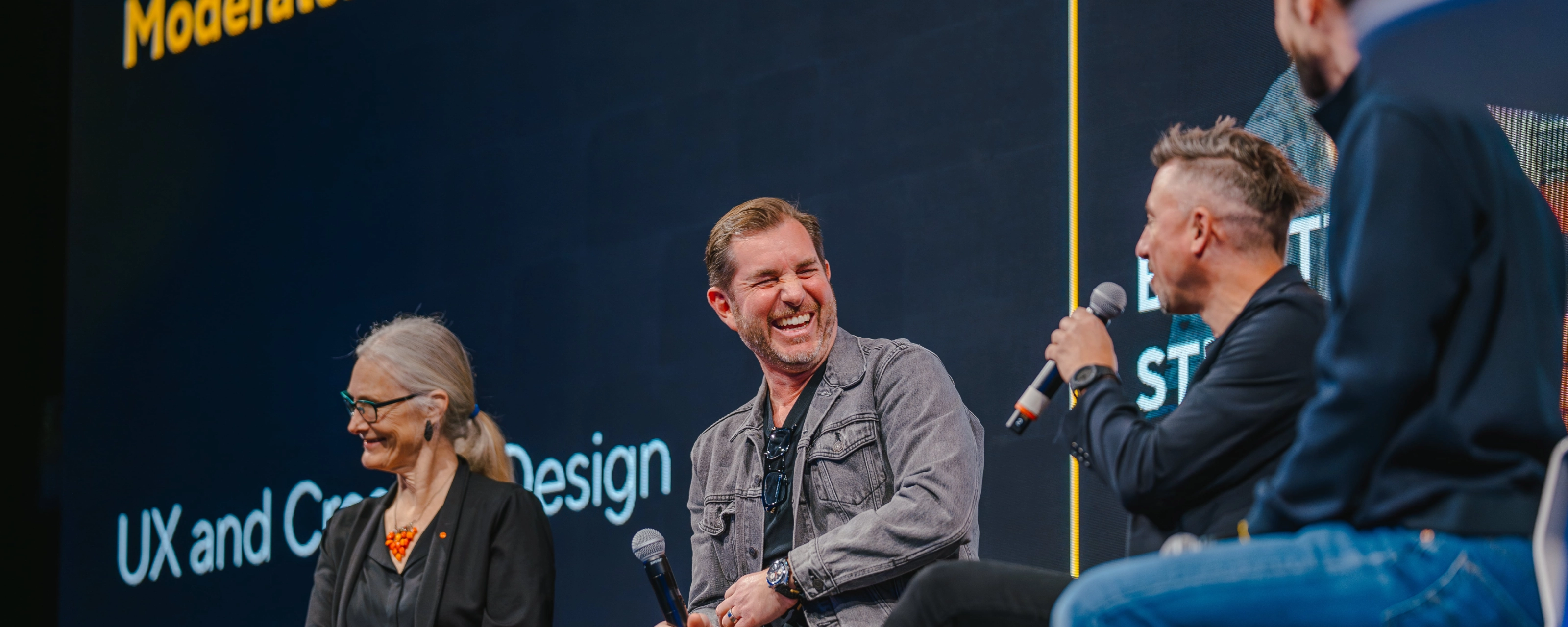 Three people with microphones laugh during a panel discussion on stage.