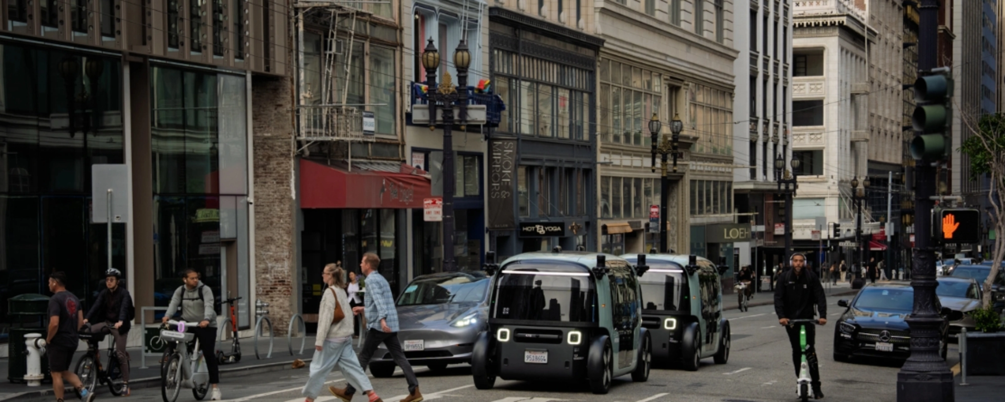 Zoox autonomous shuttle driving through a city intersection with pedestrians nearby.
