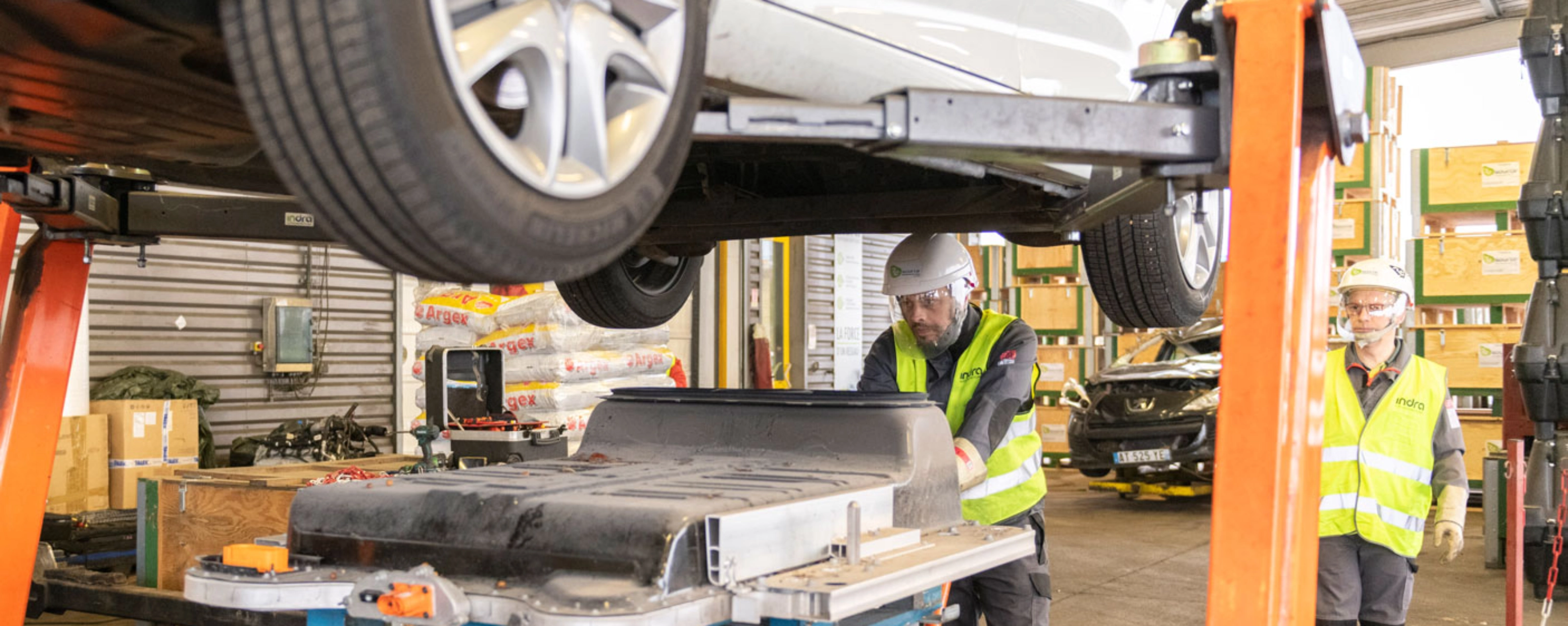 Worker in factory handling EV battery module under vehicle lift.