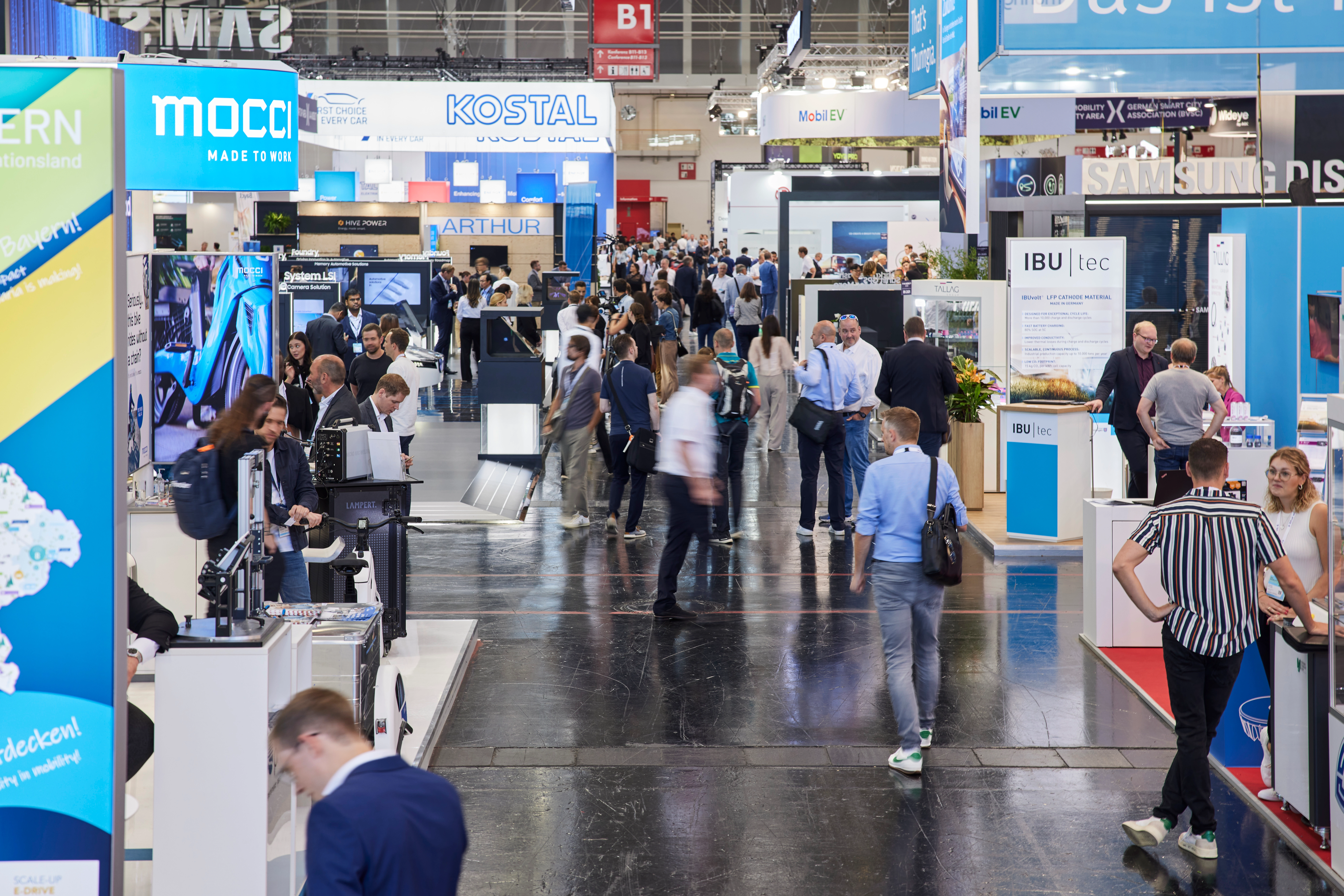 Busy trade fair hall with numerous booths, digital displays, and visitors walking around.