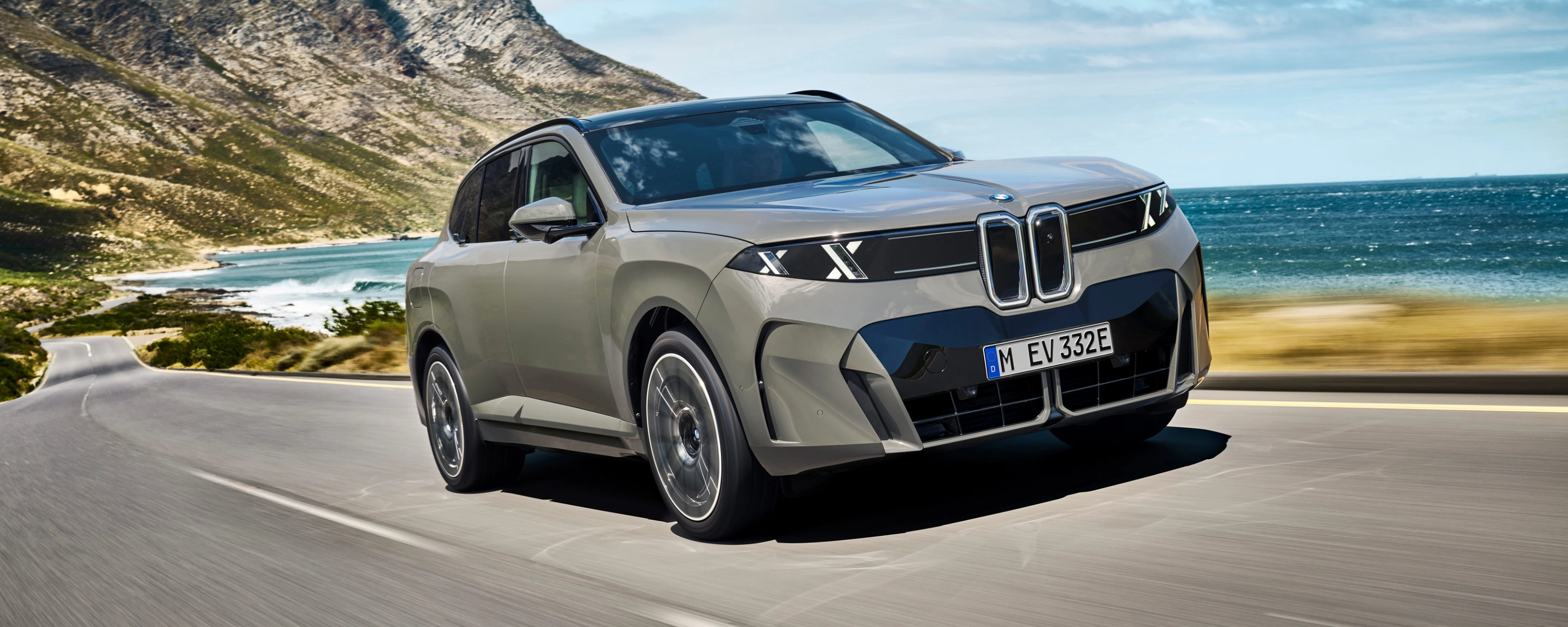 BMW SUV fährt auf einer Küstenstraße mit Bergen und Meer im Hintergrund.