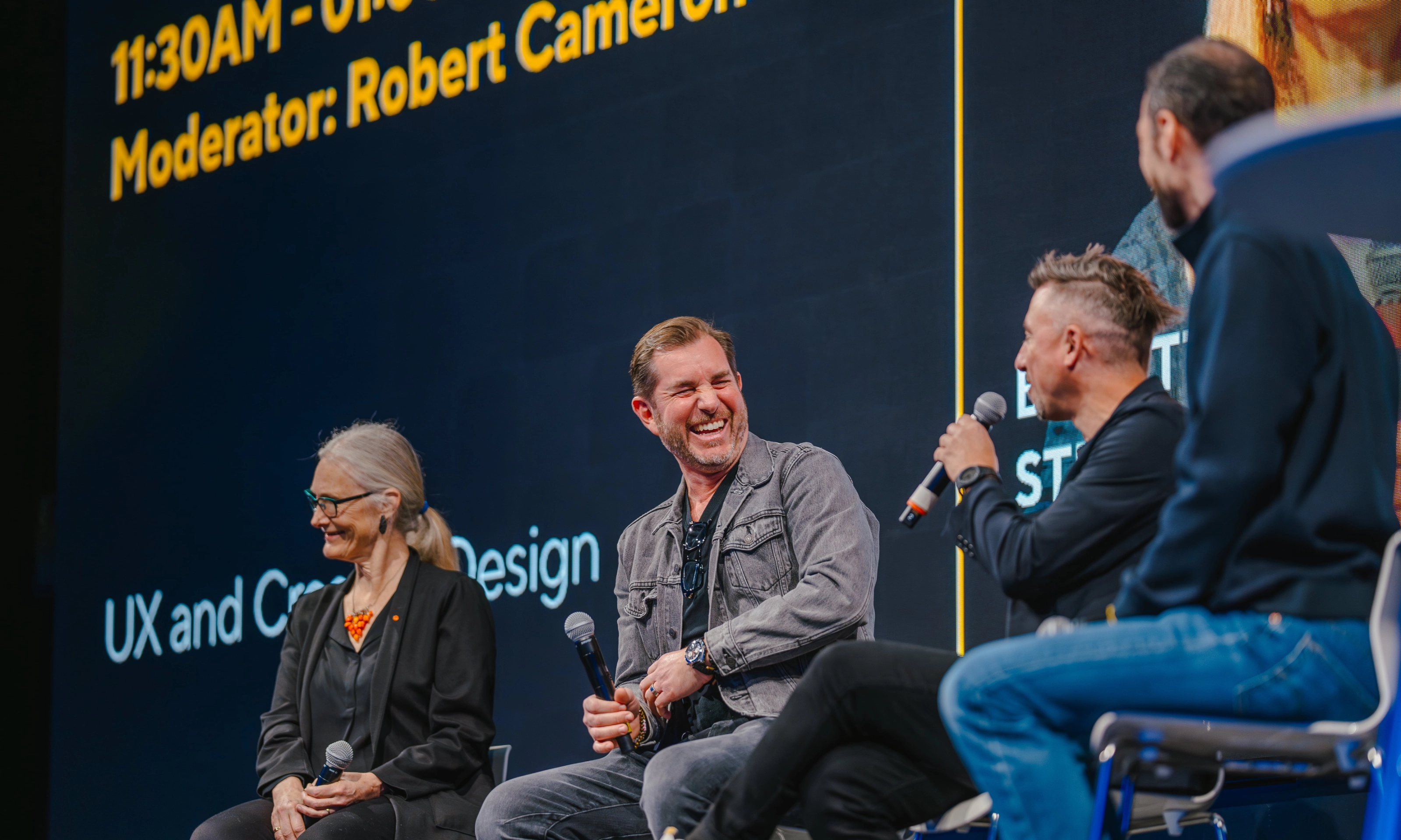 Three people with microphones laugh during a panel discussion on stage.