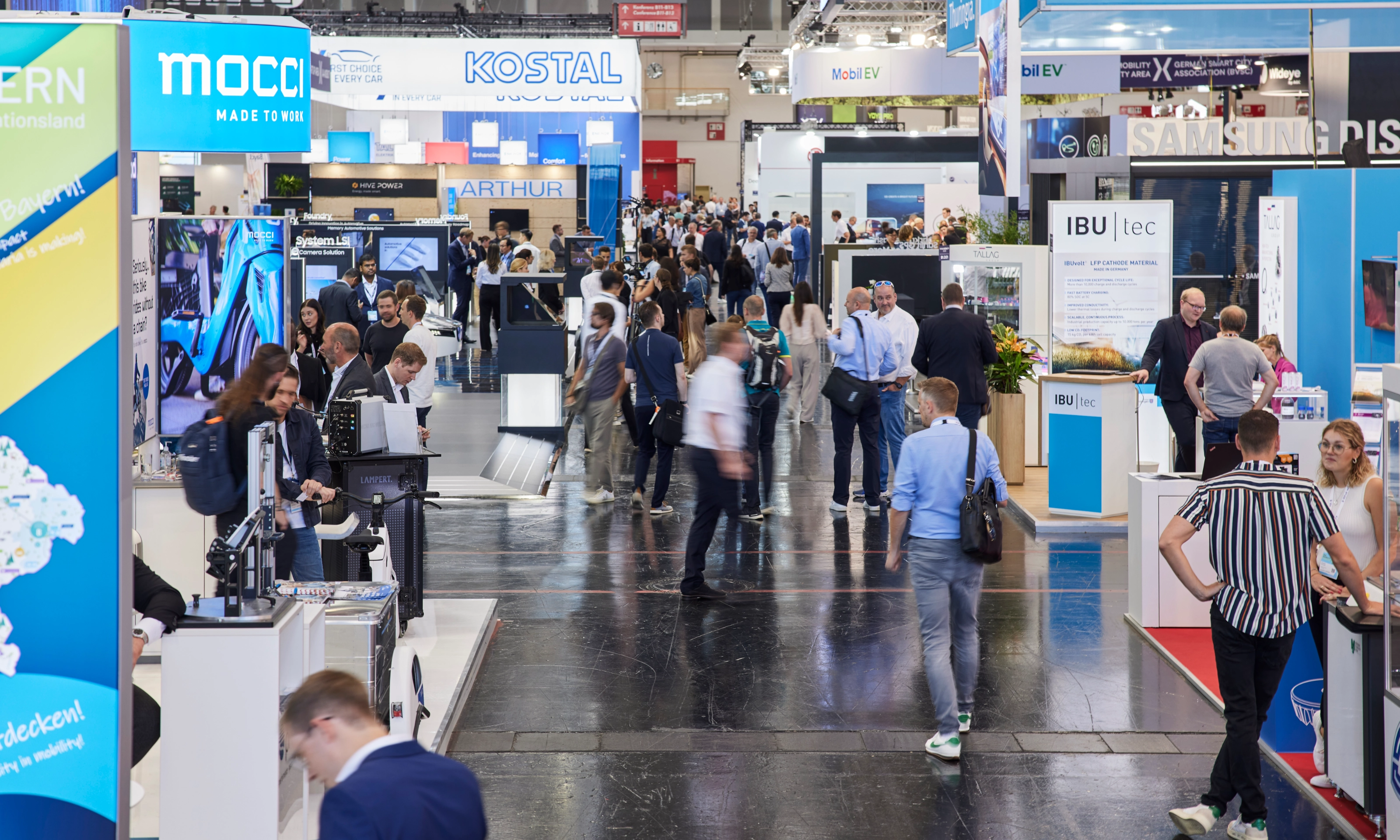 Busy trade fair hall with numerous booths, digital displays, and visitors walking around.