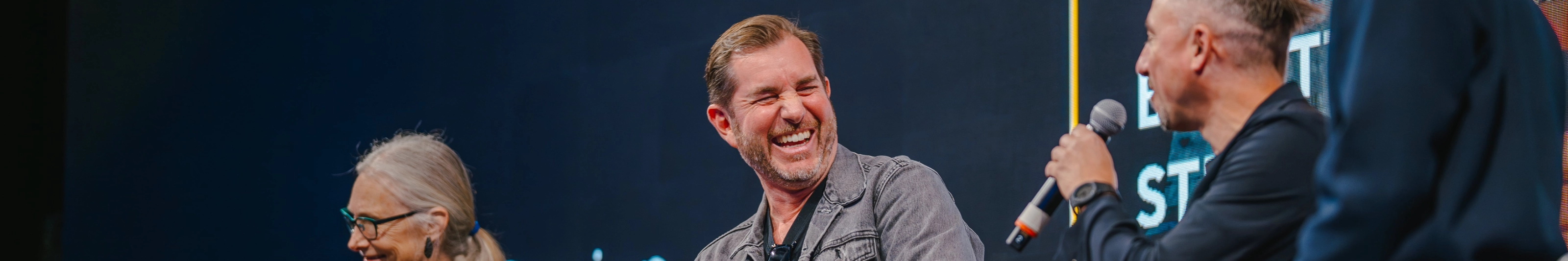 Three people with microphones laugh during a panel discussion on stage.