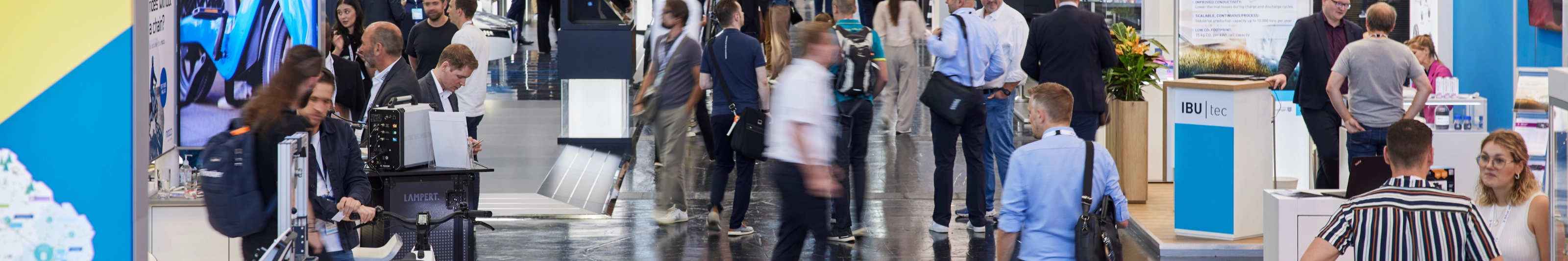 Busy trade fair hall with numerous booths, digital displays, and visitors walking around.