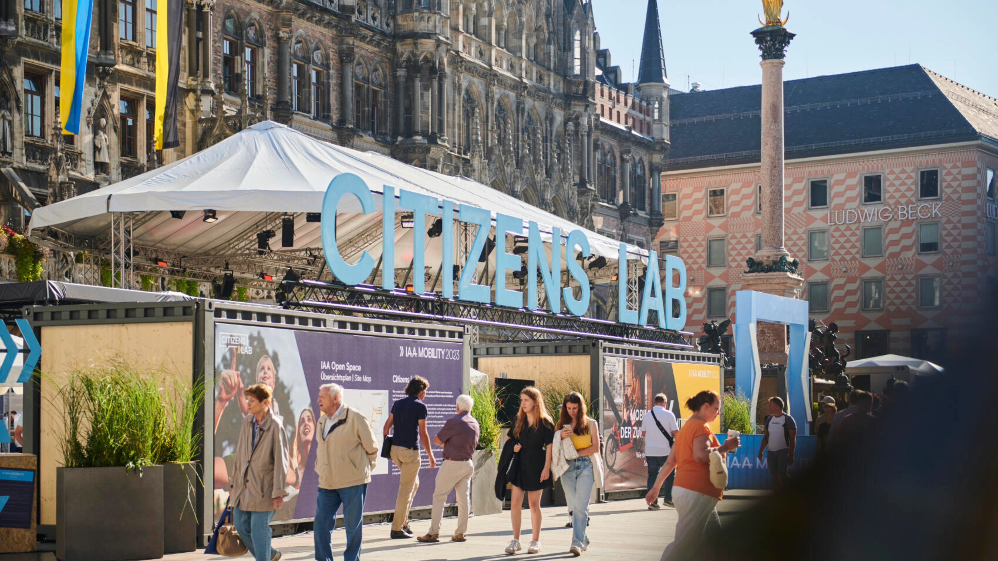 Outdoor scene at the IAA Mobility 2023 Citizens Lab in Munich, showing a large container installation with signage and visitors walking by in front of historic buildings.